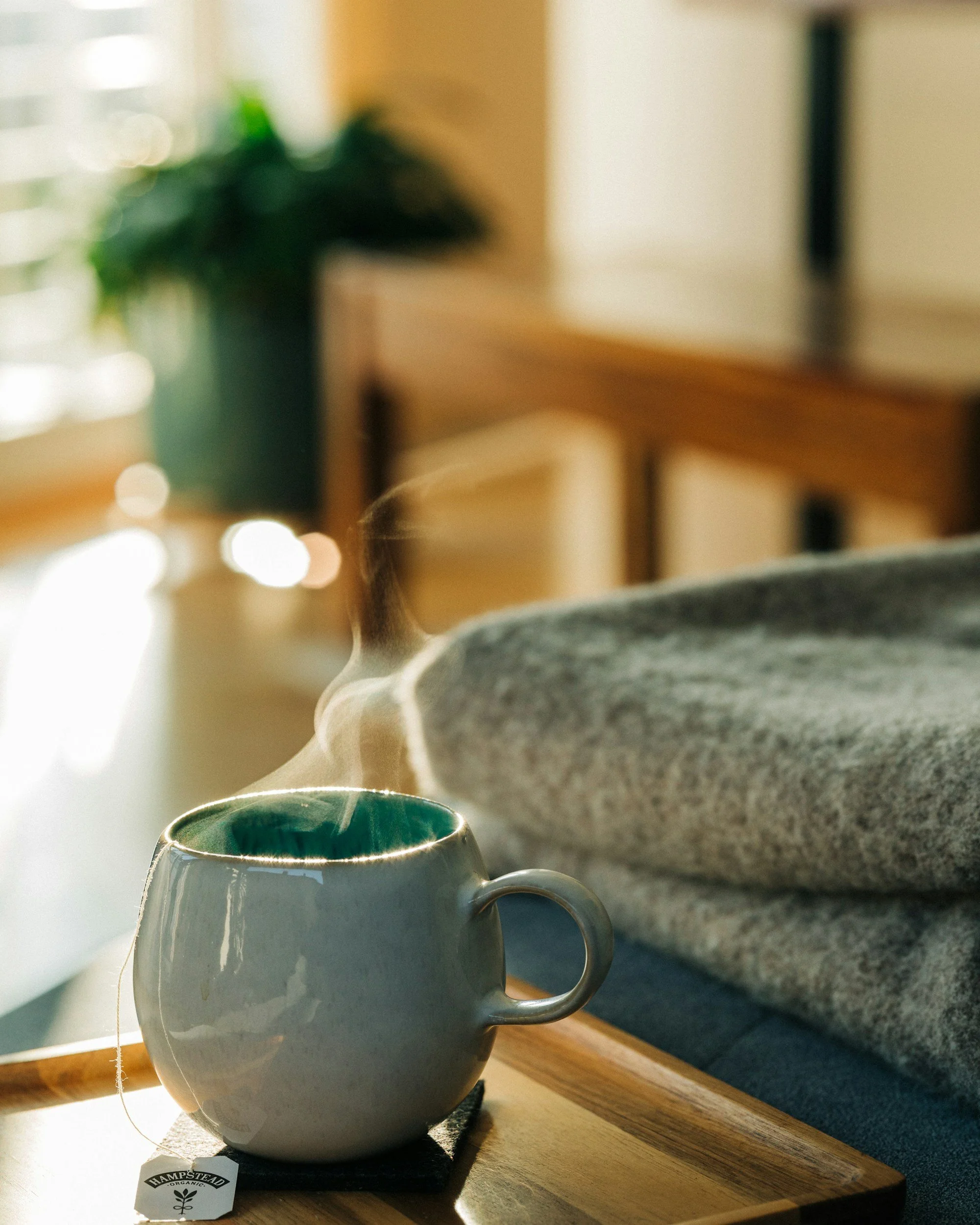A white ceramic mug with steam rising from it, placed on a wooden surface with sunlight streaming in. A tea bag with a tag that reads 'Hampstead Organic' is inside the mug. In the background, there is a granite countertop and a blurred potted plant.