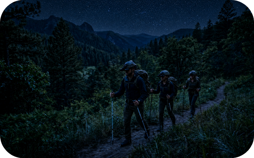 Three hikers with backpacks and trekking poles walking on a trail through a forested mountain landscape at night under a starry sky.