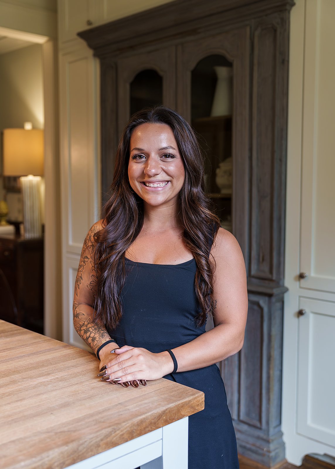 A woman with long dark hair, wearing a black sleeveless top, smiling and sitting at a wooden counter in a kitchen, with wooden cabinets and a lamp in the background.