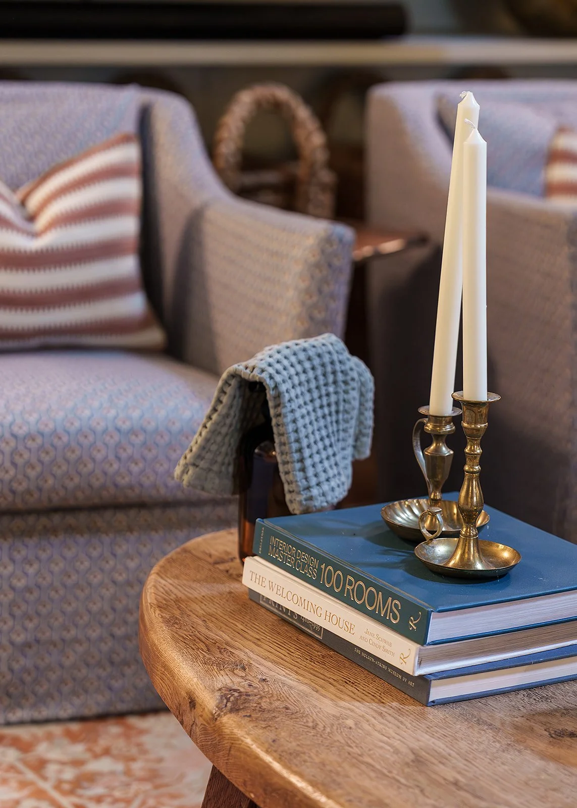 Close-up of a rustic wooden table with stacked books, including titles on interior design and welcoming houses, with a brass candlestick holder and tall white candles, in a cozy living room with patterned upholstered sofas and pillows in the background.