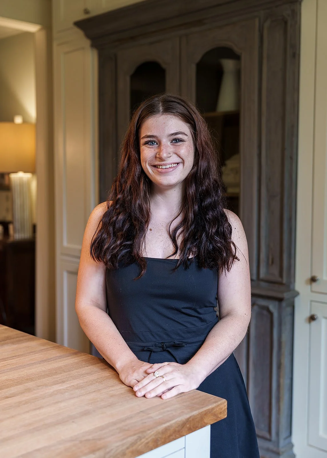 A young woman with long, wavy, dark red hair standing in a kitchen, smiling at the camera, wearing a black sleeveless top and matching black apron.