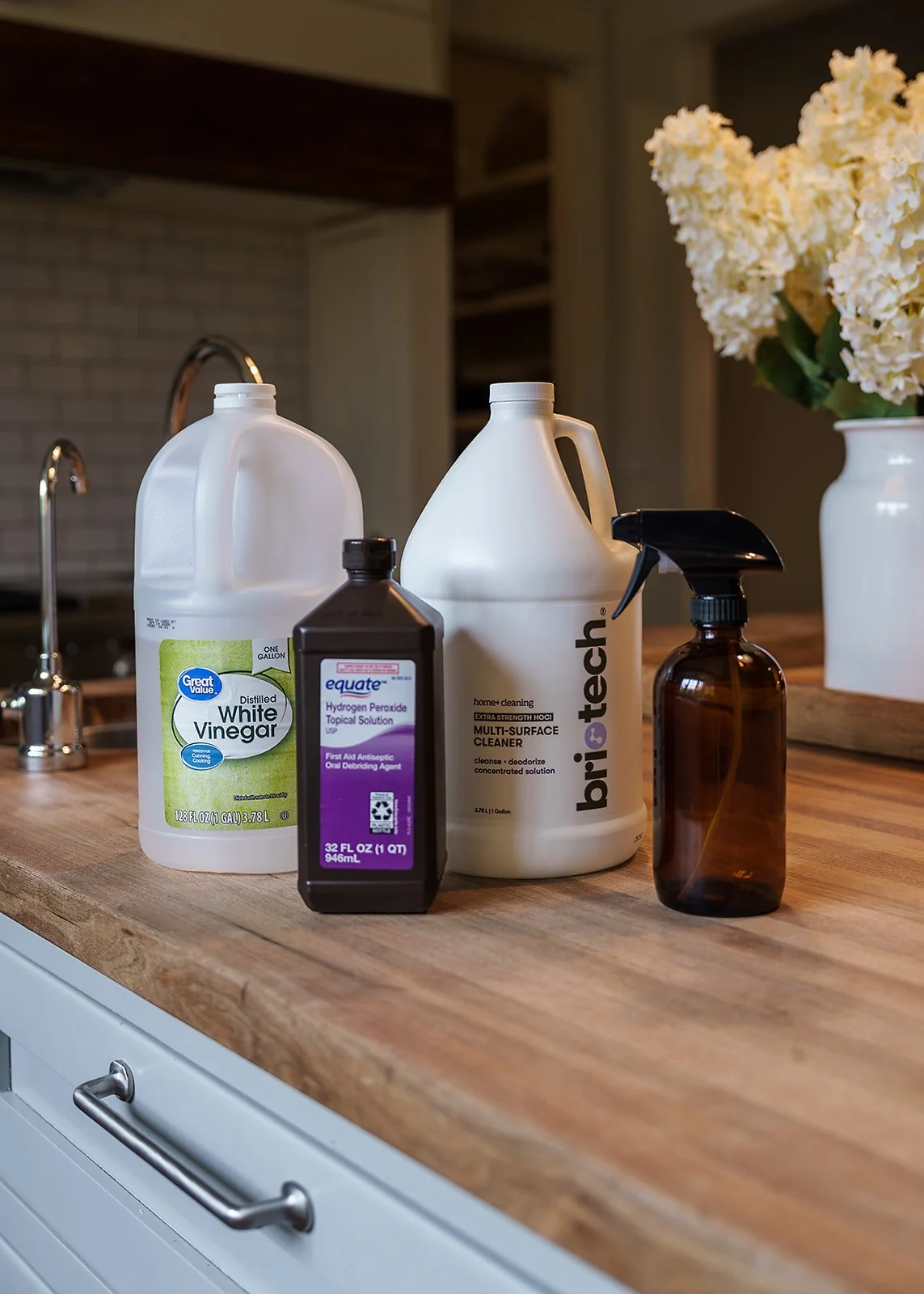Various cleaning supplies on a wooden kitchen countertop, including white vinegar, hydrogen peroxide, multi-surface cleaner, and an amber spray bottle, near a vase of white flowers.