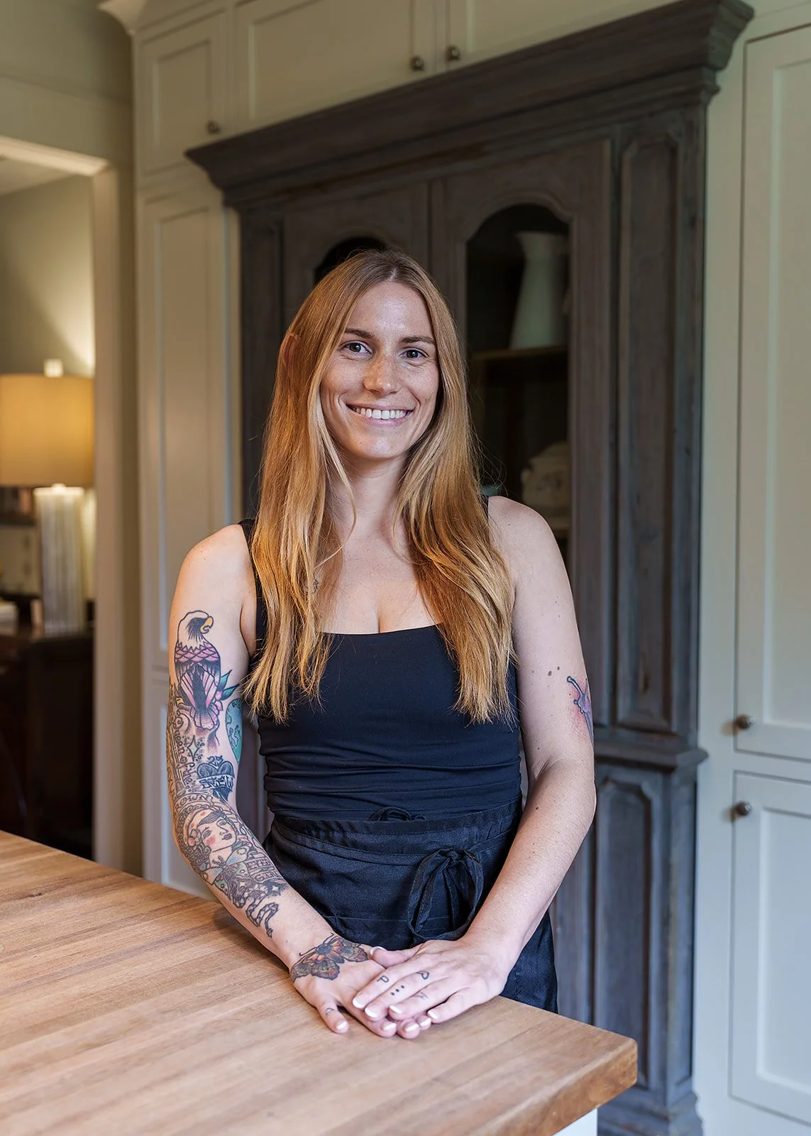 A smiling woman with long red hair, tattoos on both arms, wearing a black sleeveless top and apron, standing in a kitchen with wooden and white cabinetry, and a warm light in the background.