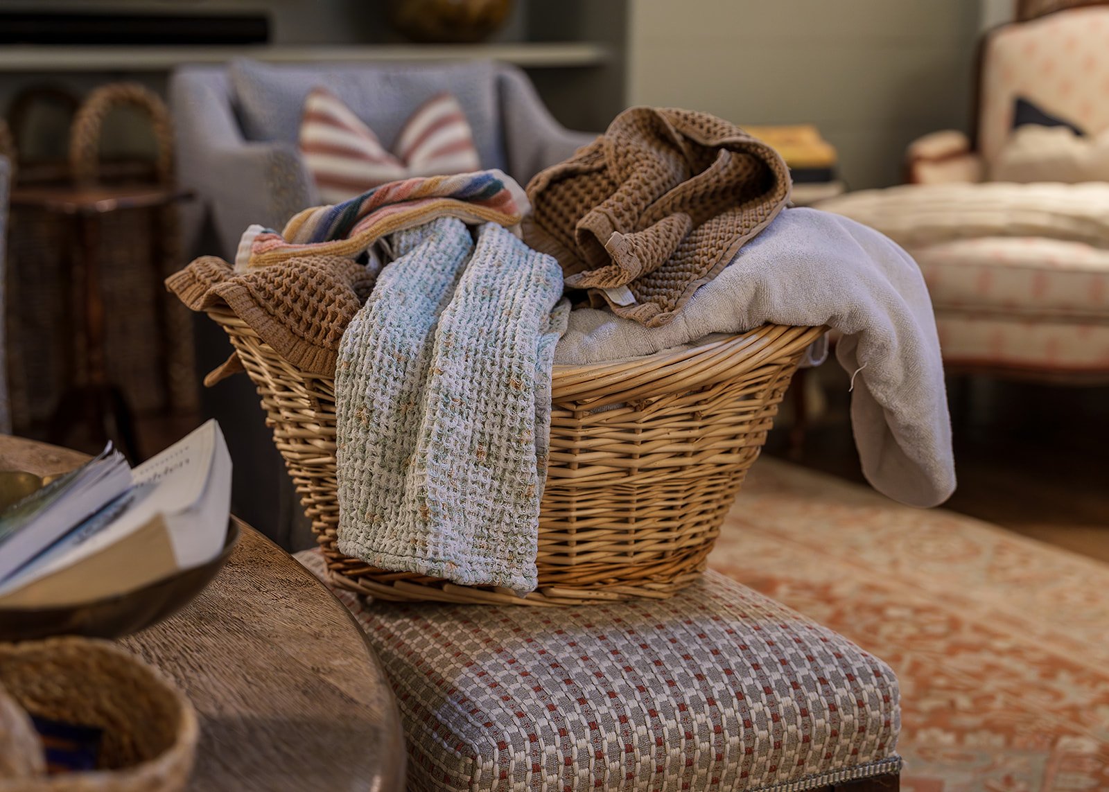 A woven basket filled with various folded towels and cloths in a cozy living room with armchairs, a side table, and carpeted floor.