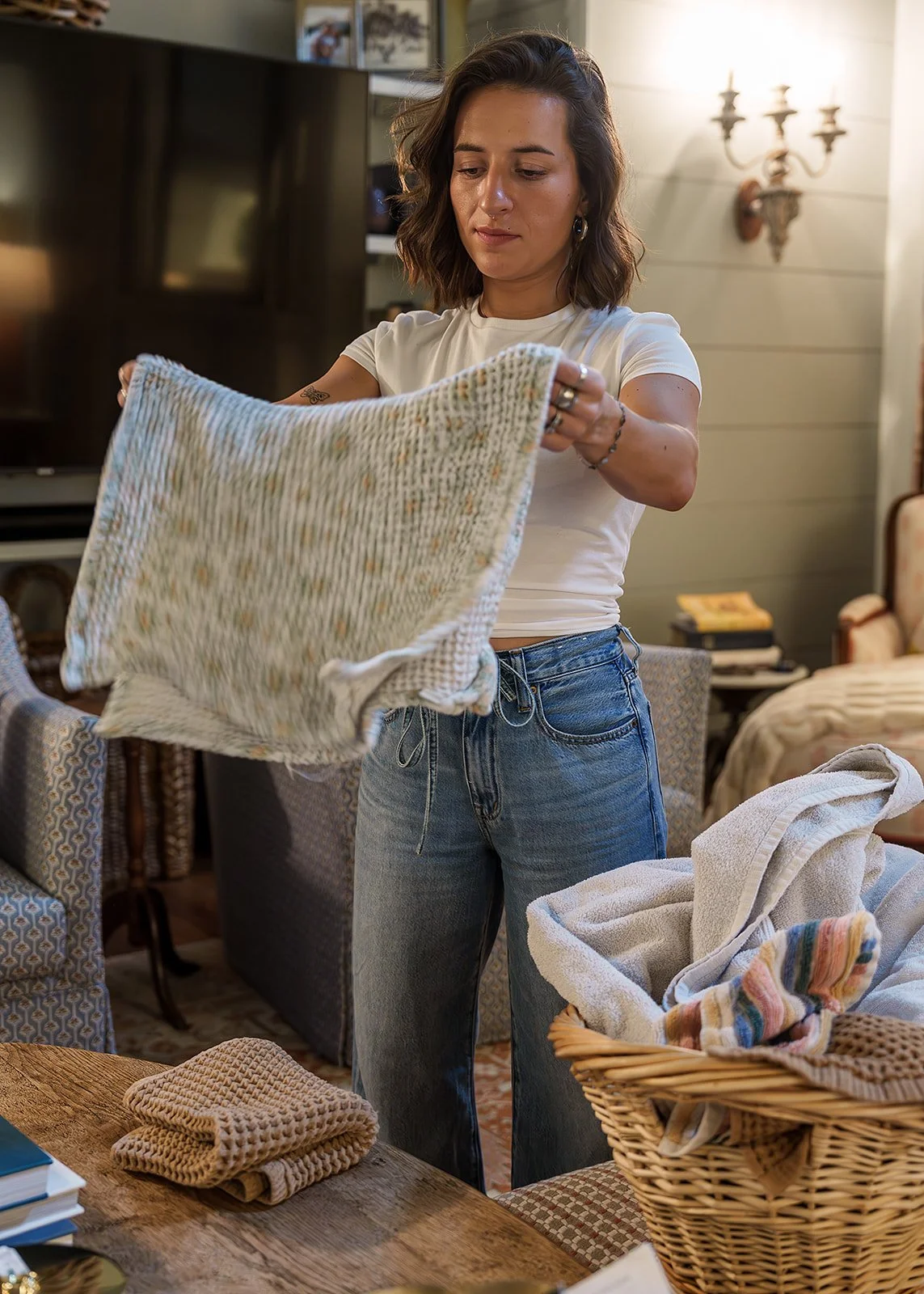 A woman in a white t-shirt and jeans folding or handling a blanket or throw blanket in a cozy, well-lit living room.