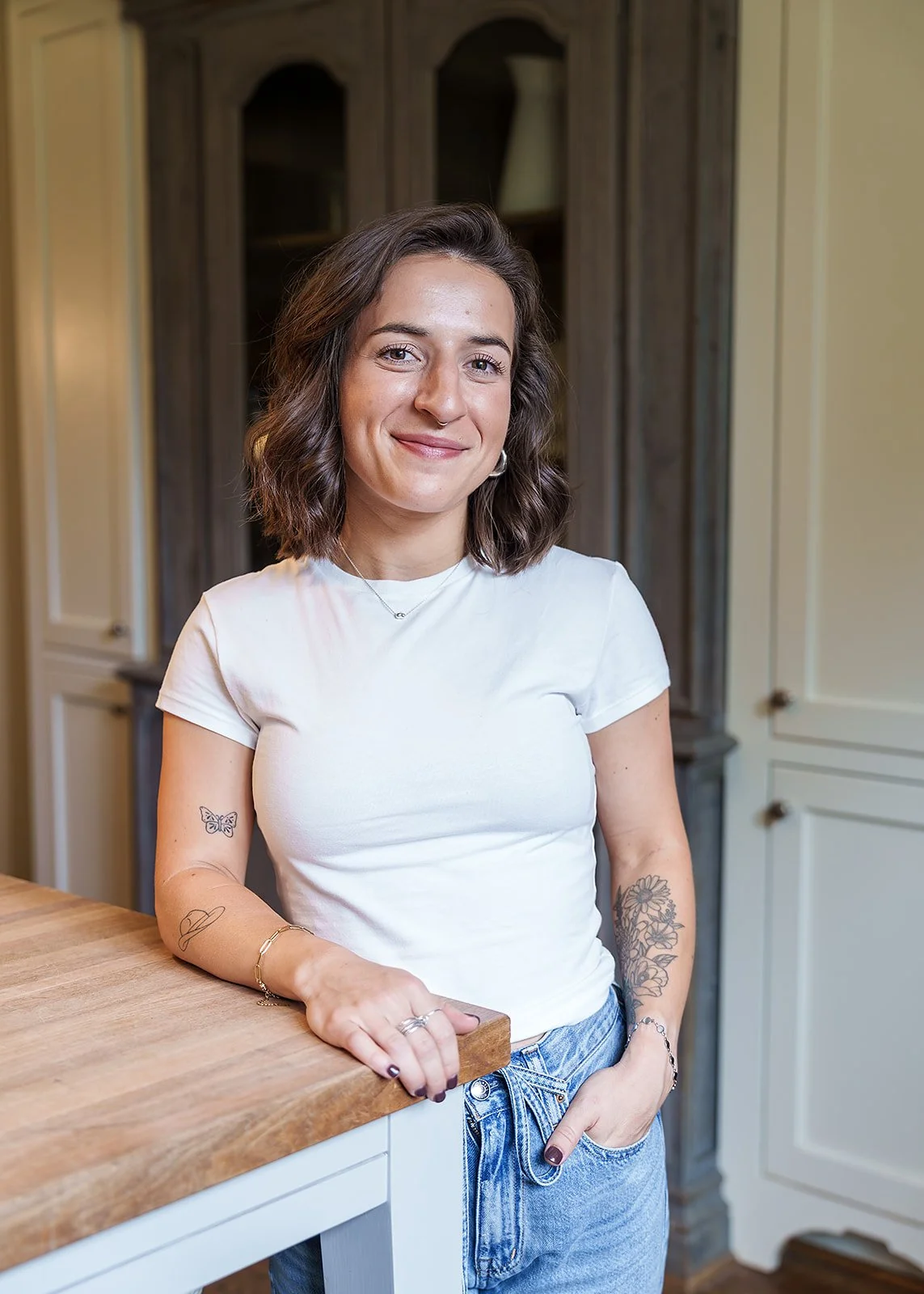 Young woman with short wavy brown hair, wearing a white t-shirt and jeans, standing in a kitchen with cabinets and a wooden countertop.