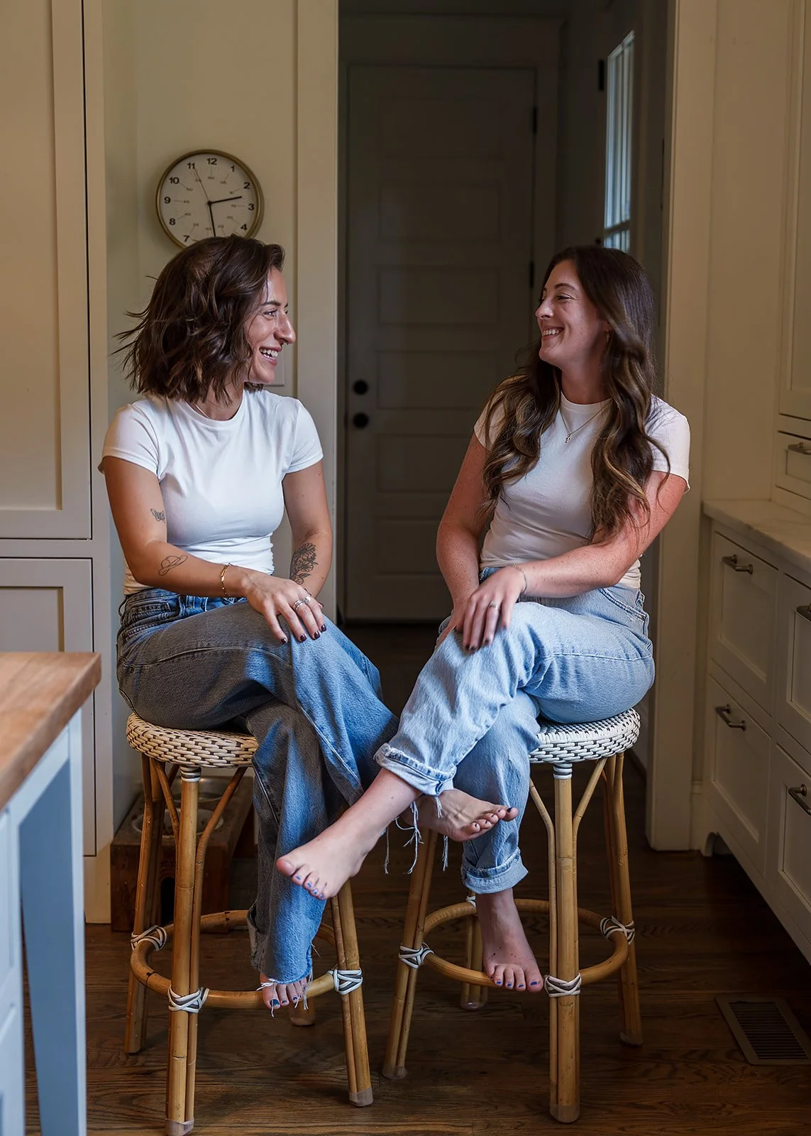 Two women sitting on bar stools in a kitchen, smiling and laughing at each other, wearing white T-shirts and jeans.