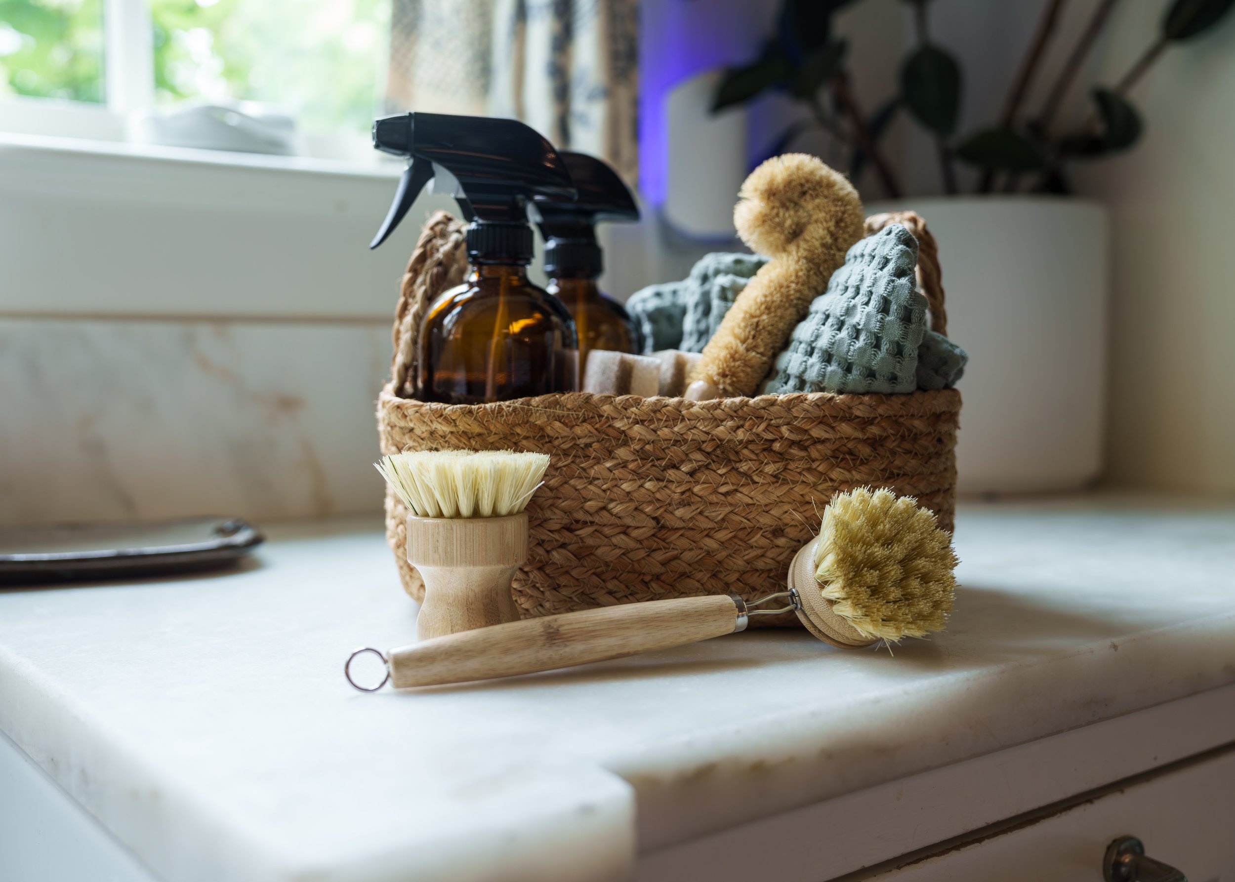 A woven basket containing cleaning supplies such as spray bottles, dishcloths, and a plush toy, placed on a white surface in front of a window with natural light.