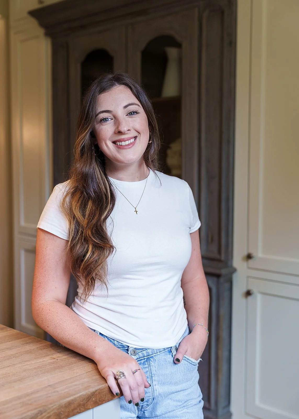 Smiling woman with long wavy brown hair wearing a white t-shirt, jeans, a cross necklace, and rings on her fingers, standing in a kitchen with wooden and cream cabinetry.