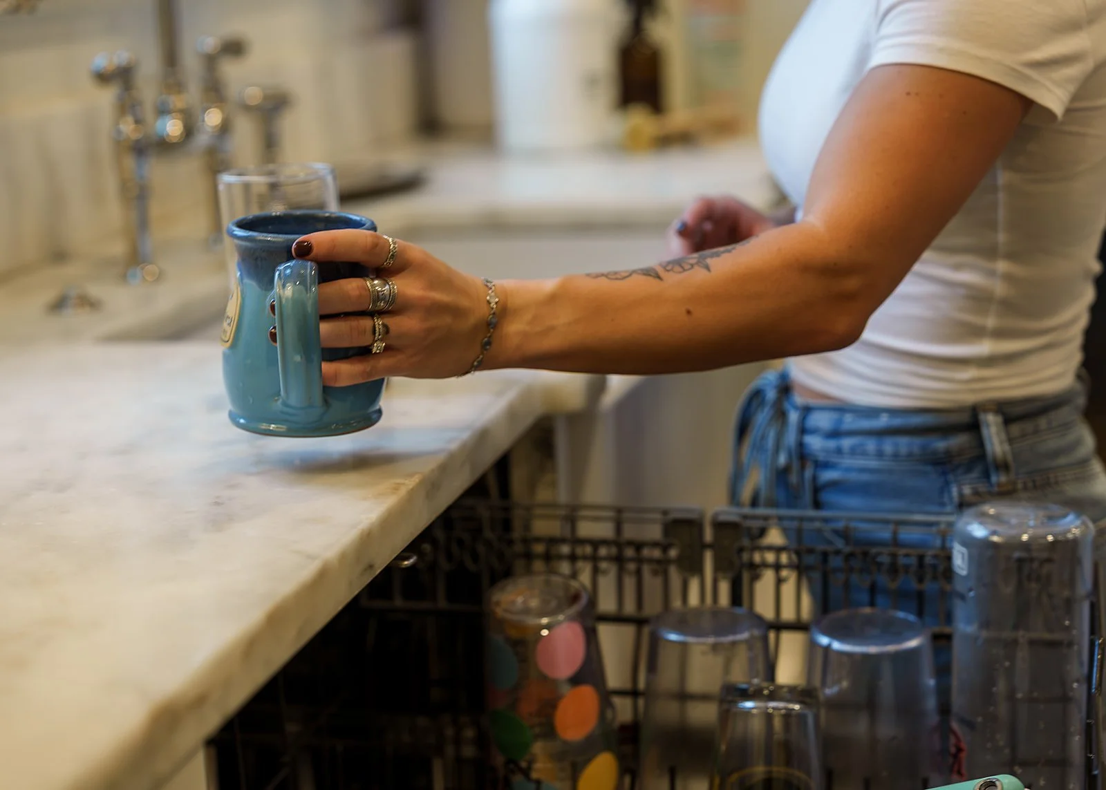 Person holding a blue coffee mug in a kitchen, wearing a white t-shirt and blue jeans, with visible tattoos and rings on their fingers.