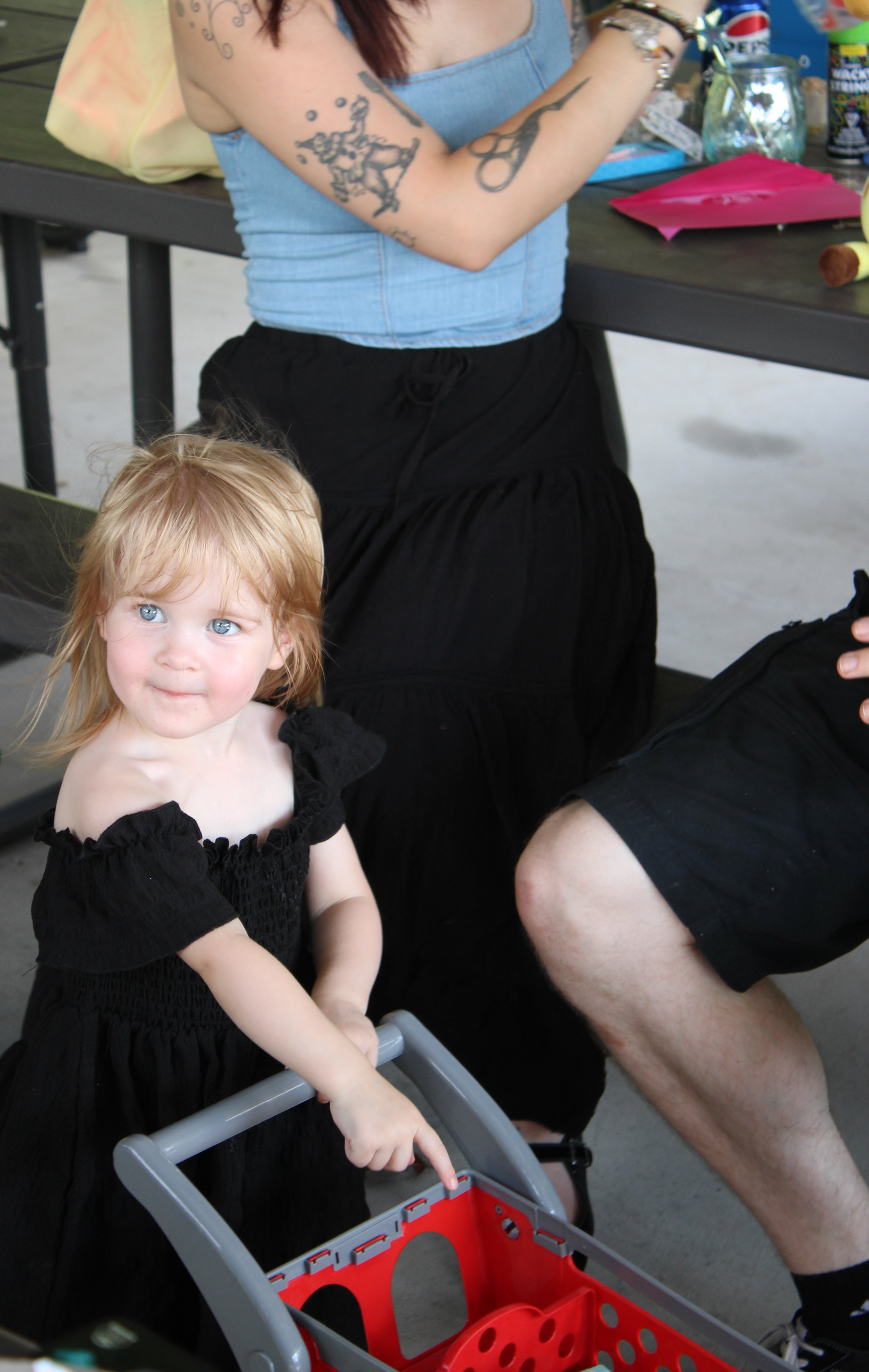 A young girl with red hair and blue eyes wearing a black dress with ruffle sleeves standing beside a red shopping cart. She is holding the cart's handle and looking slightly to the side. An adult woman, partially visible, sits nearby wearing a light 