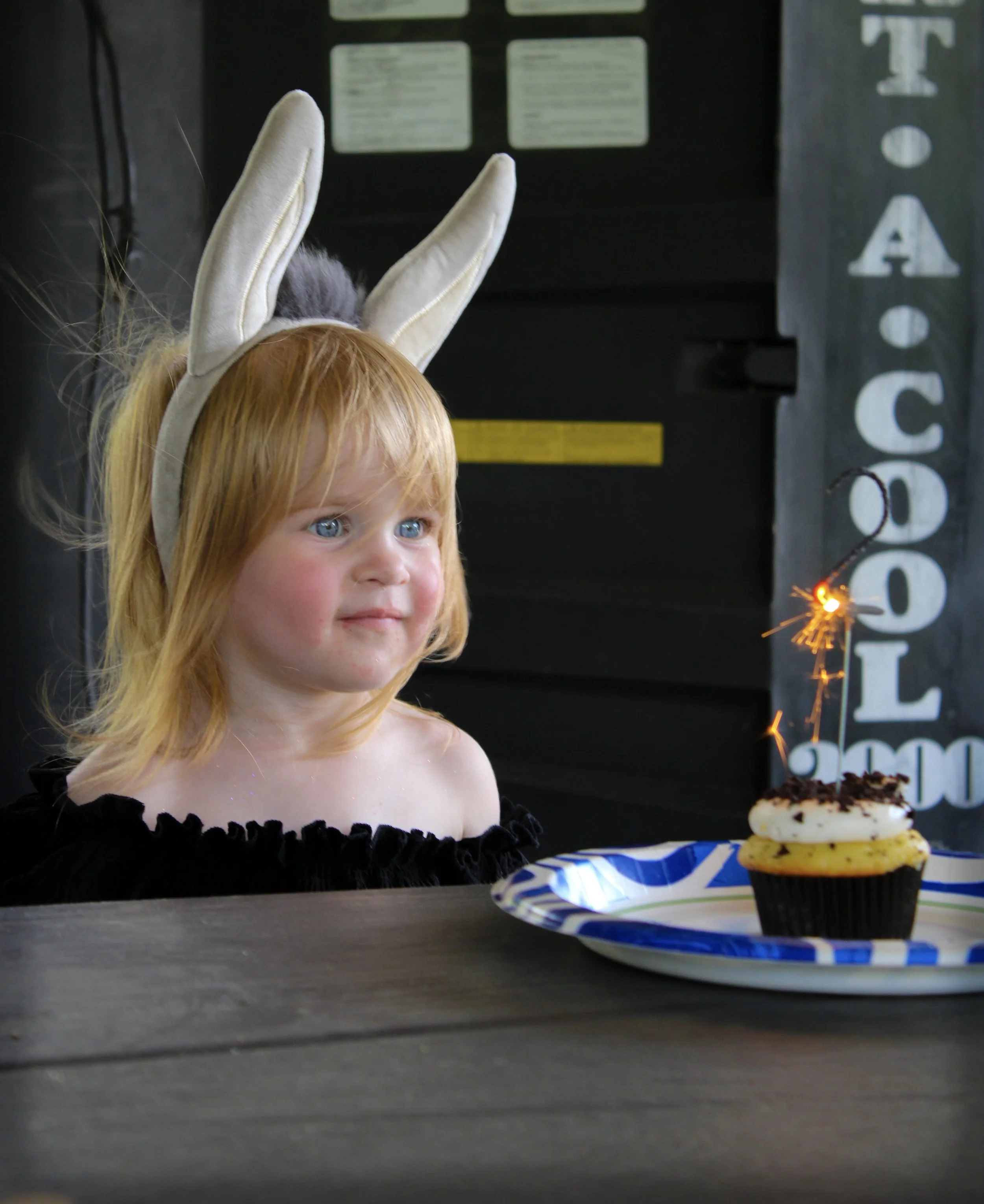Close-up of a young girl with red hair, blue eyes, and bunny ears headband, sitting in front of a birthday cake with a sparkler, in a room with black walls and white letters.