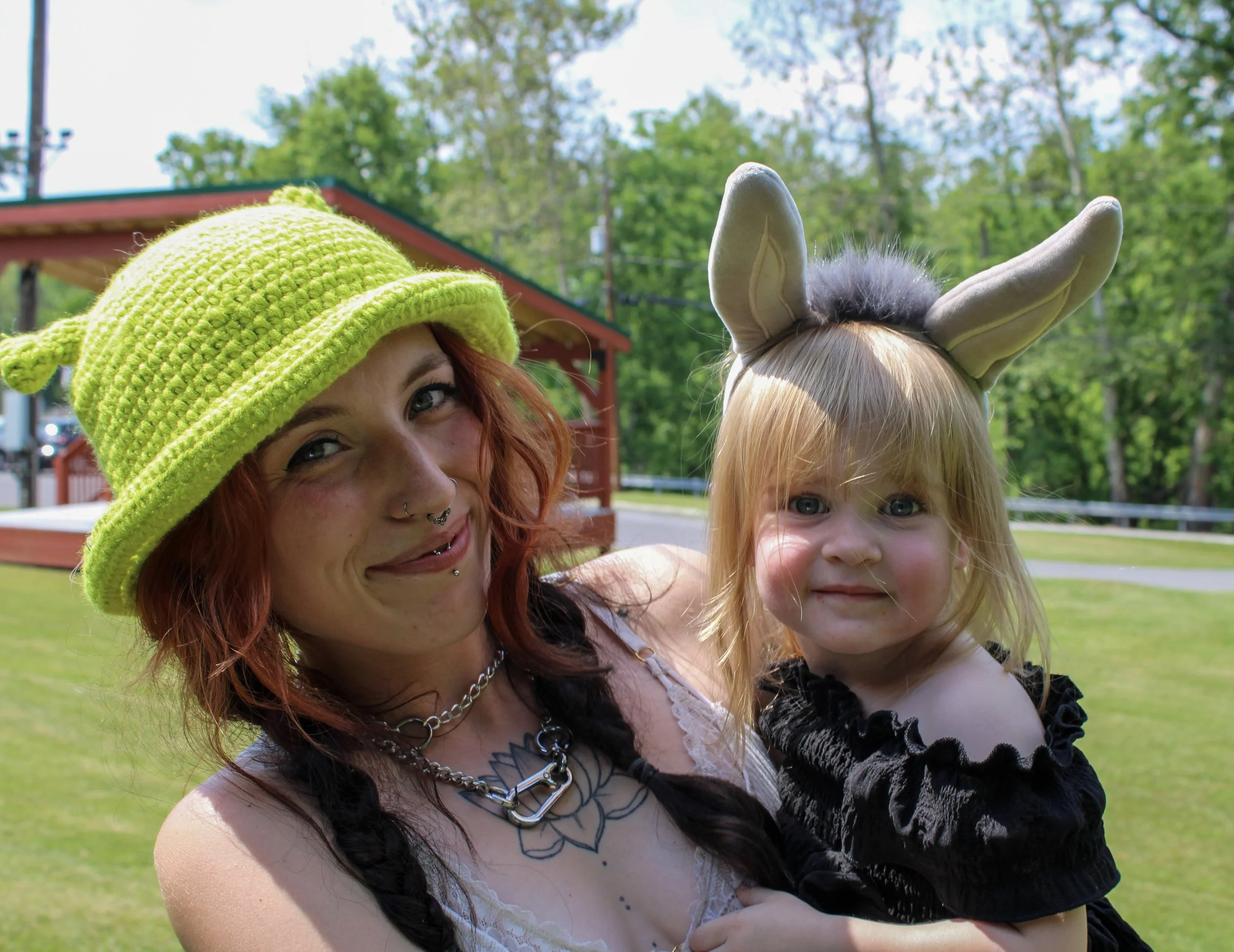A woman with red hair wearing a lime green knit hat with animal ears, and a tattoo on her shoulder, smiling with a young girl with blond hair and blue eyes wearing a gray headband with animal ears, outdoors on a sunny day with trees and a building in