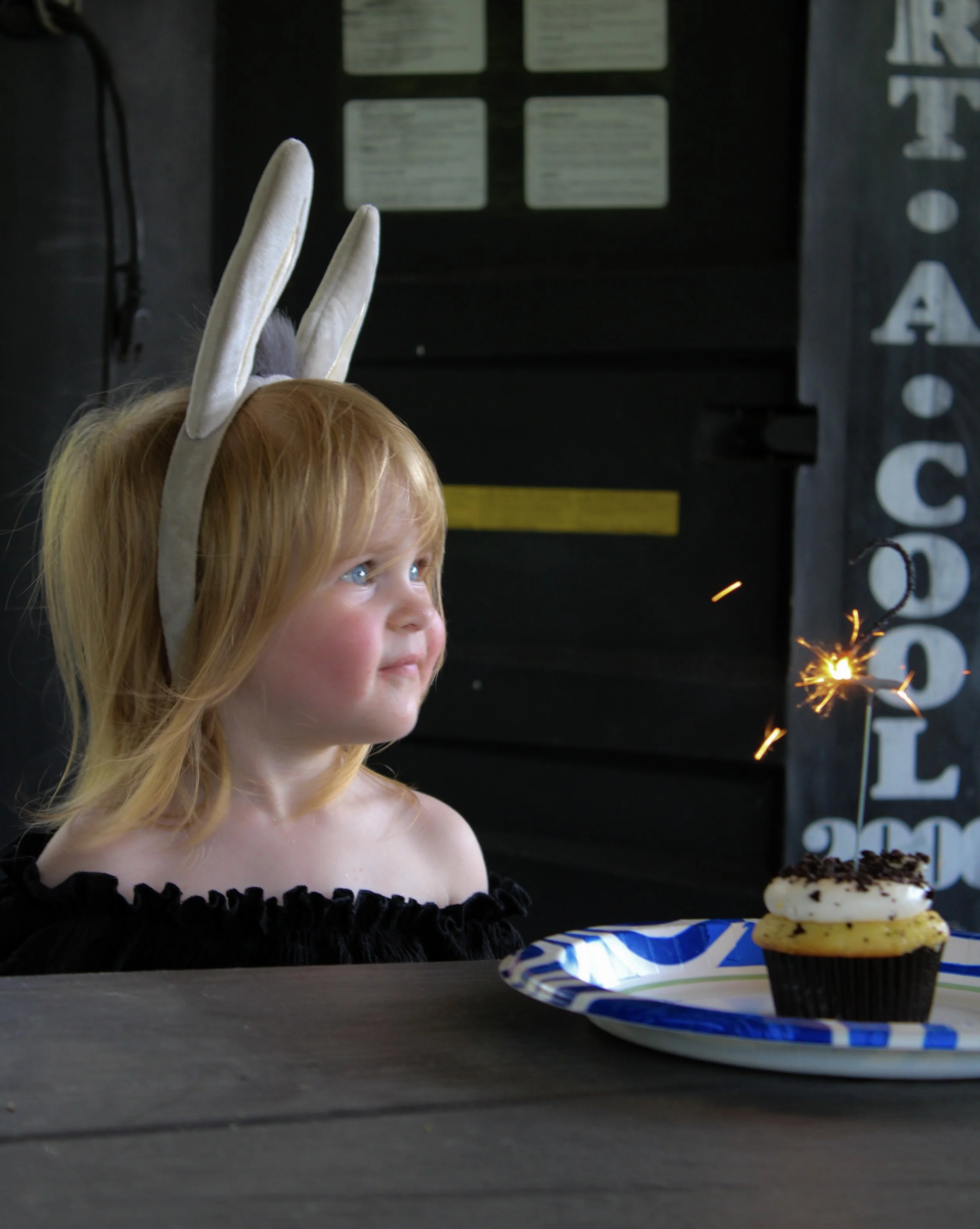 A young girl with red hair wearing bunny ears headband, black dress with ruffled neckline, celebrating her birthday with a chocolate cupcake topped with white frosting and sprinkles on a blue and white striped plate. In the background, there is a cho