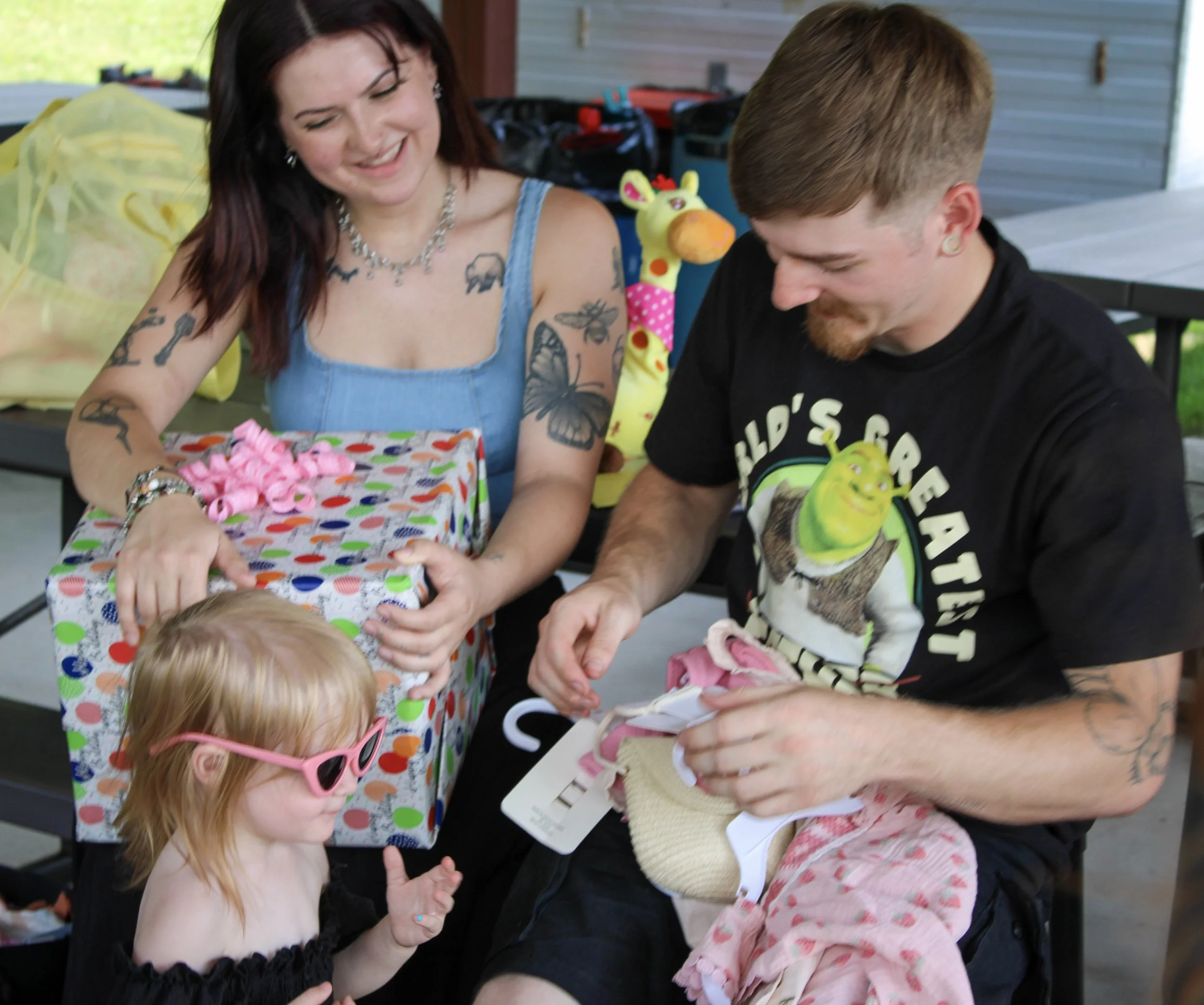 A woman with tattoos and dark red hair smiling and opening a gift, surrounded by children, at an outdoor gathering.