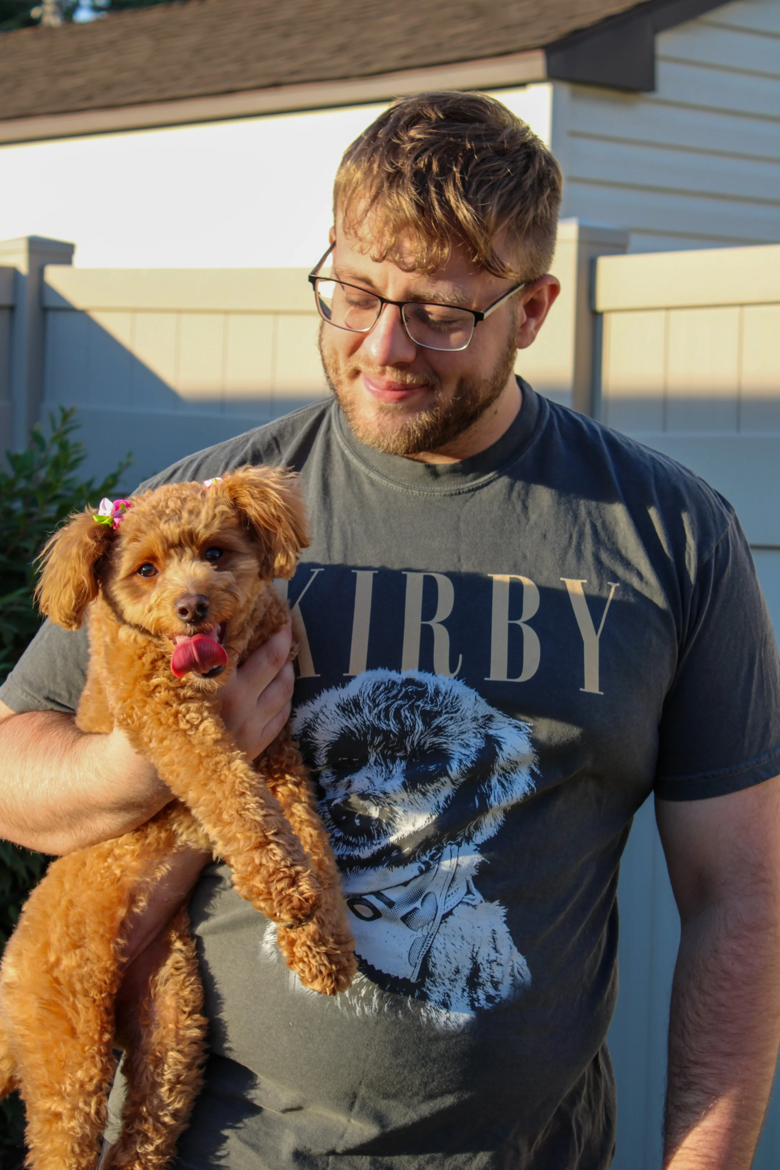 A man with glasses holding a small curly-haired dog with a pink flower in its hair, standing outdoors near a white fence and house.