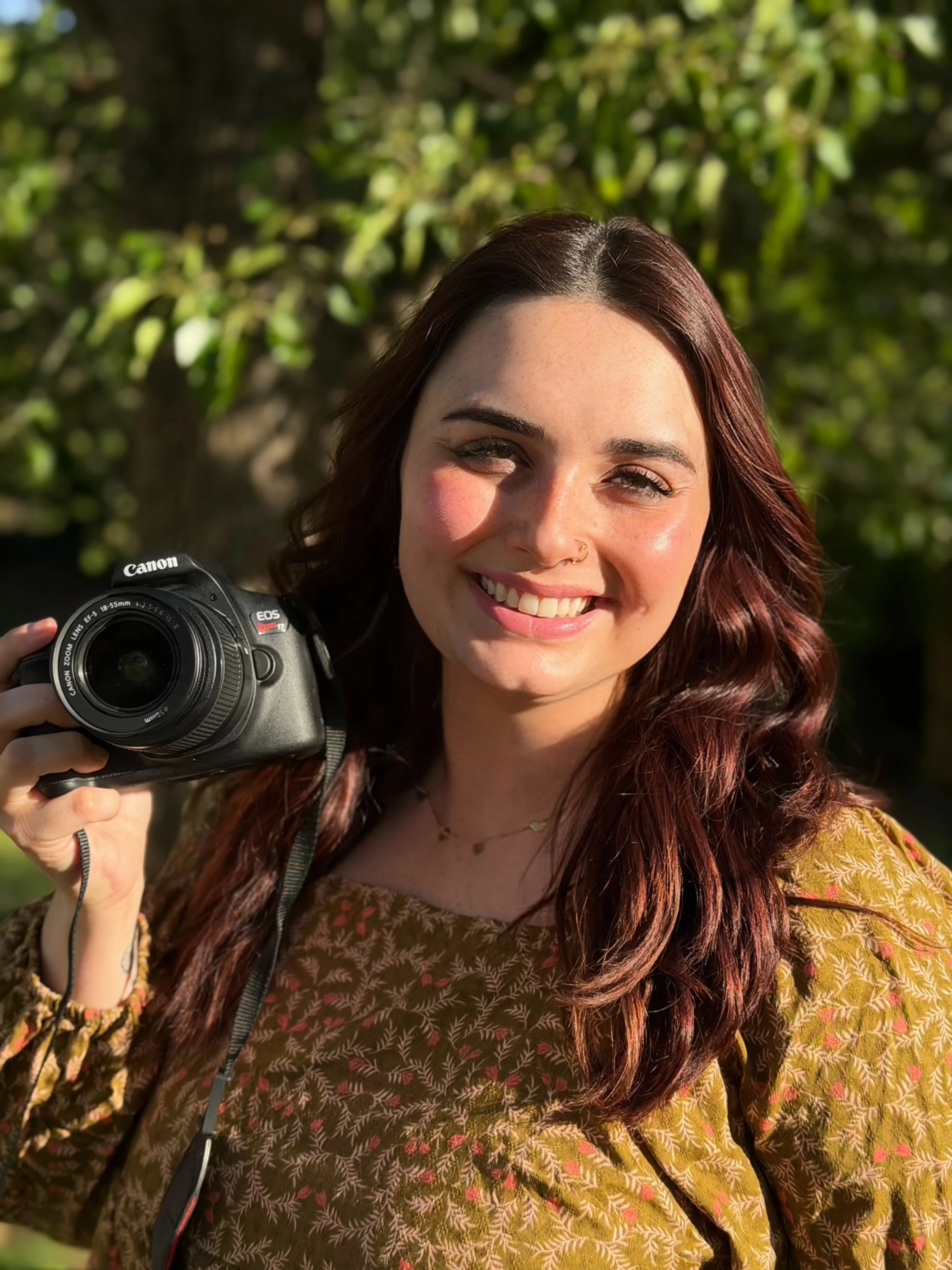 A young woman with wavy red hair smiling while holding a Canon camera outdoors, with green foliage in the background.