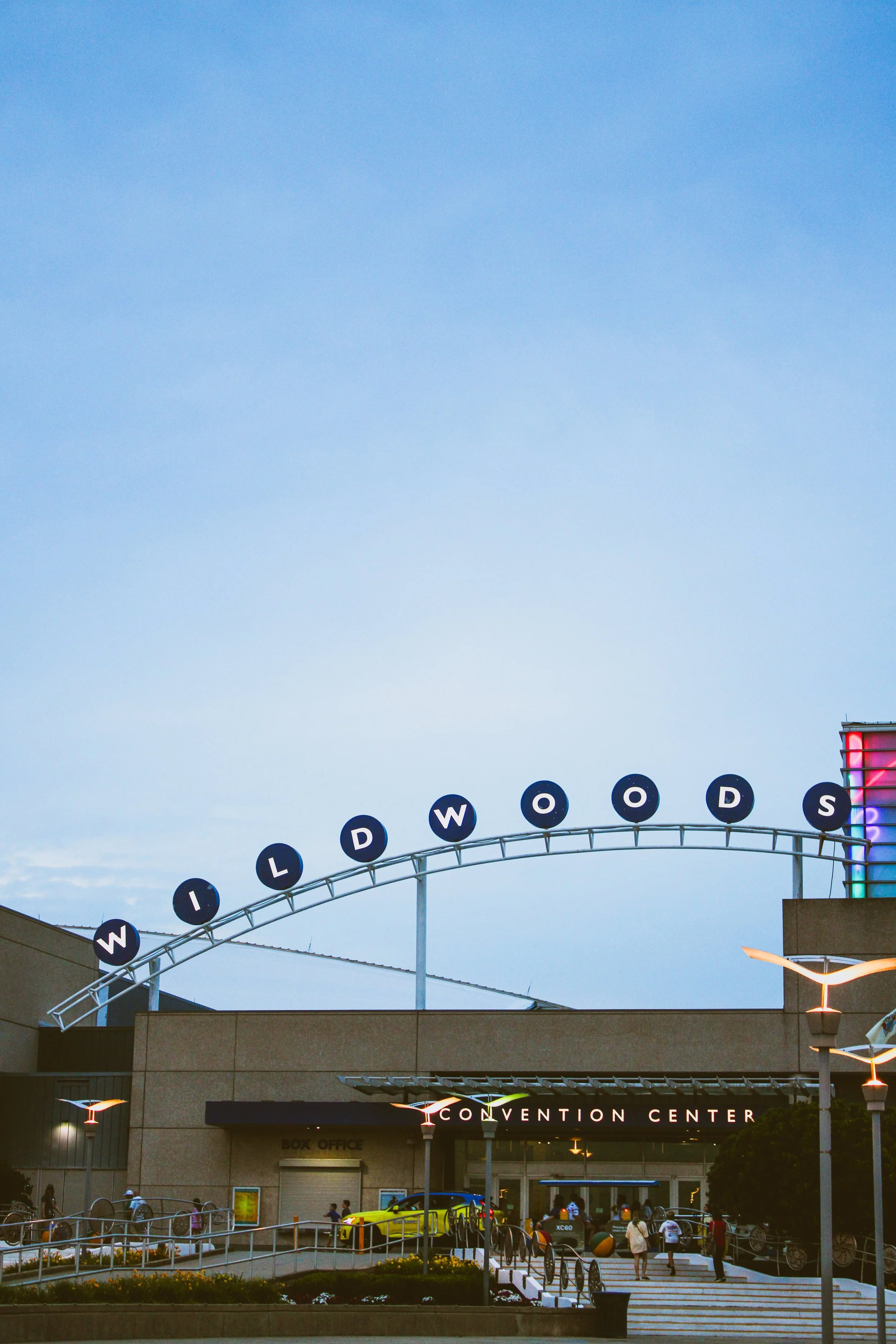 Exterior view of a convention center with a curved sign displaying the words 'WILL RODGERS.' The building has a modern design, including a glass tower with colorful lighting, and people are walking around the entrance.
