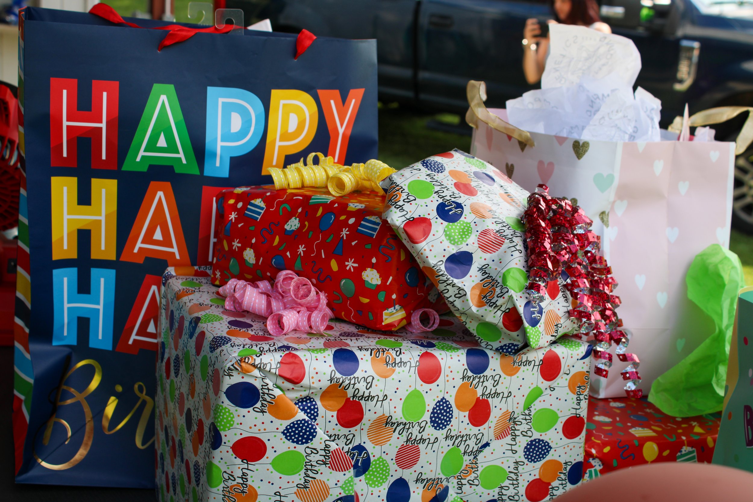 Colorful birthday presents wrapped in festive paper with ribbons and bows, with a large gift bag reading 'HAPPY BIRTHDAY' in multicolored letters in the background.