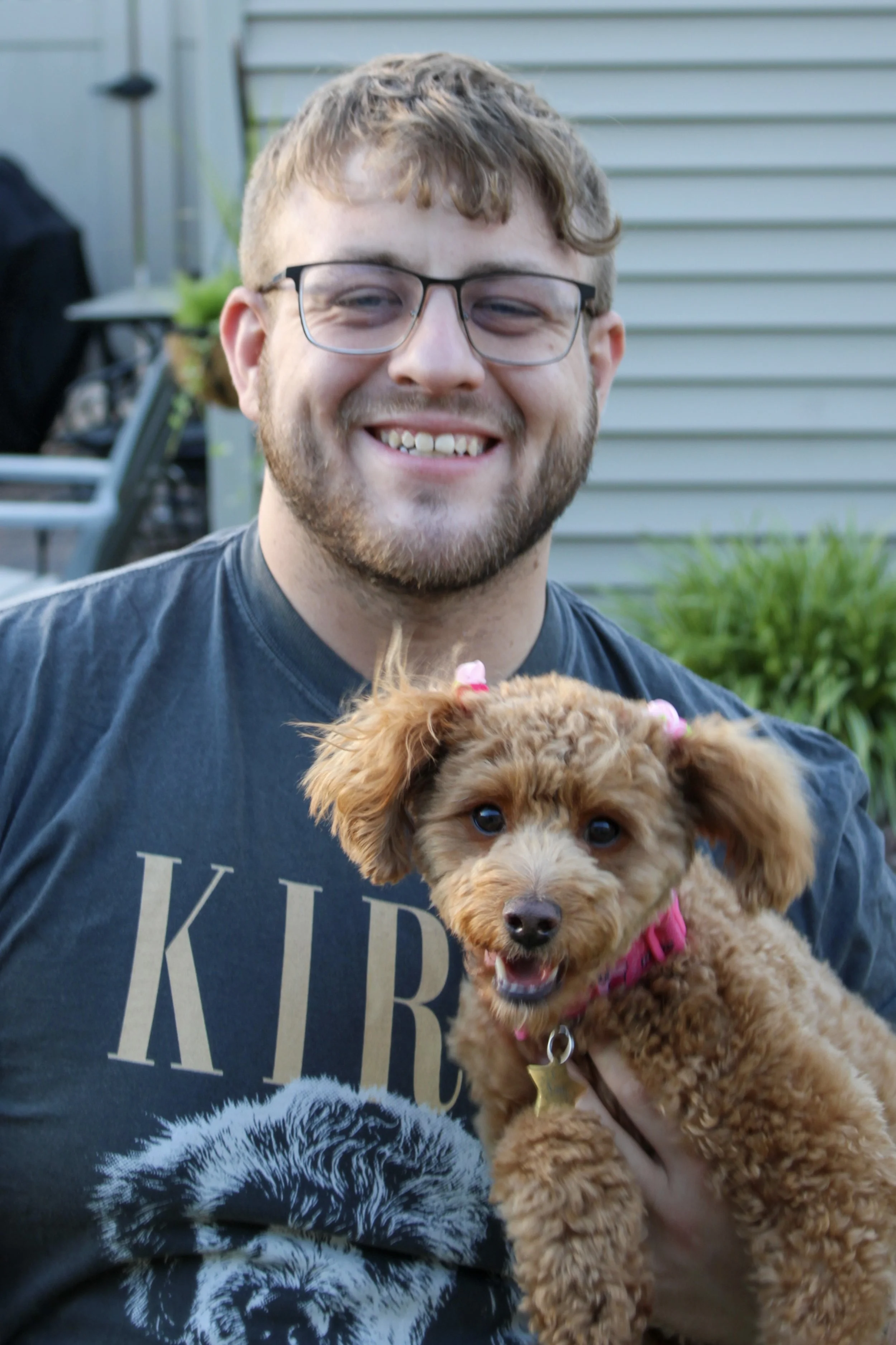 A young man with glasses smiling and holding a fluffy brown puppy outdoors against a house sidings background.