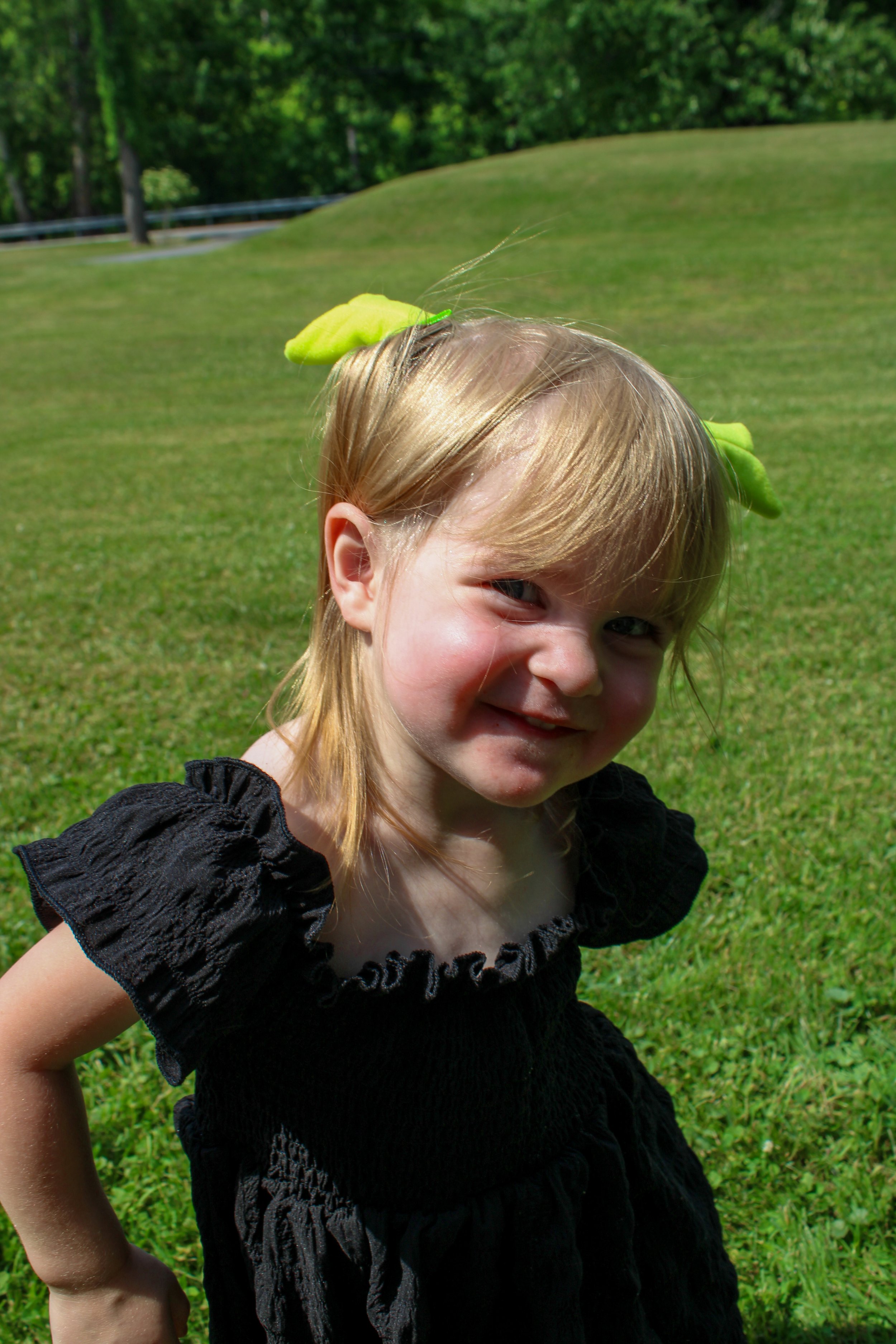 A young girl with blond hair smiling and squinting outdoors on green grass, with a large green leaf on her head.
