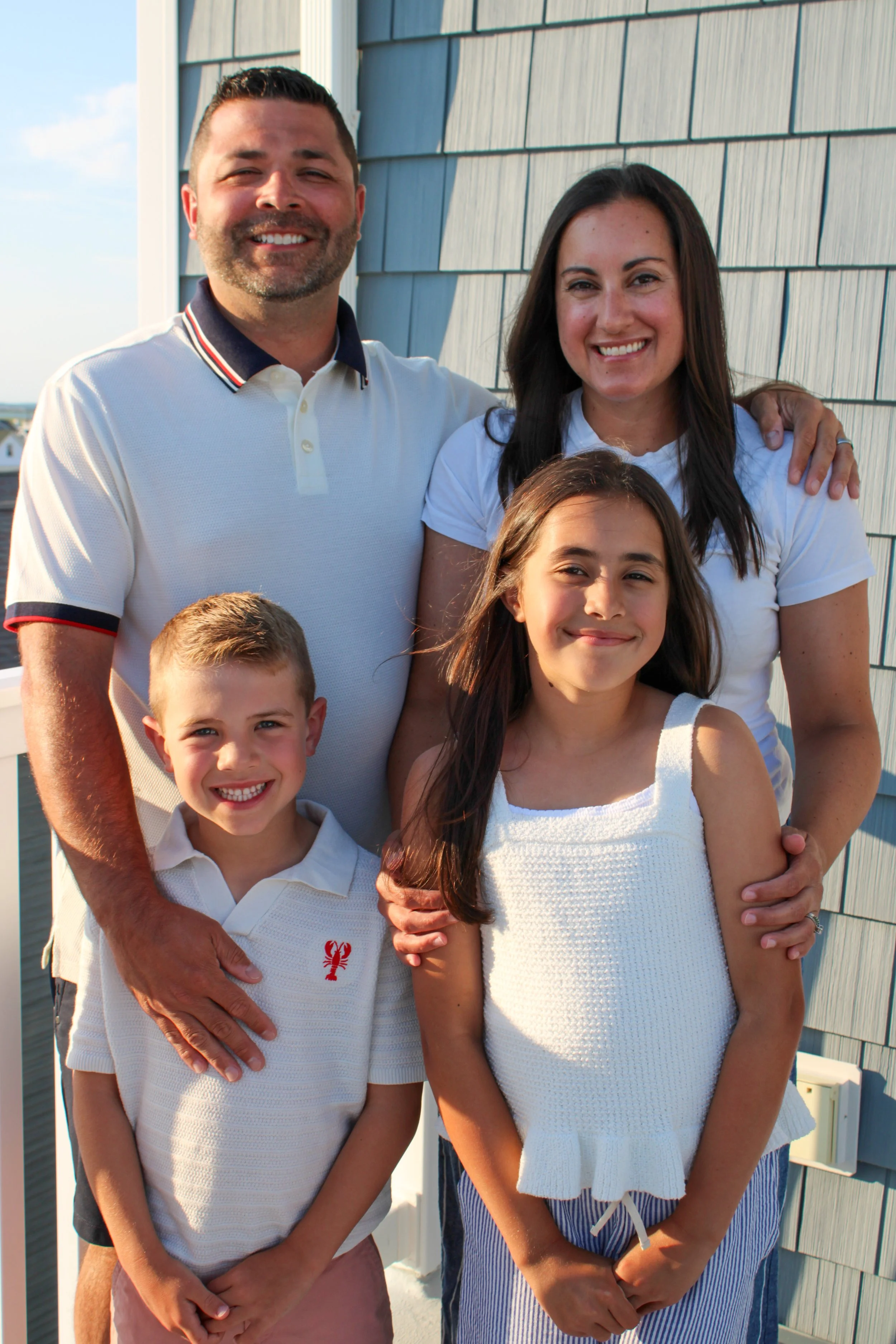 A smiling family of five posing outdoors near a house with blue siding during sunset. The group includes a man, woman, and three children, all wearing casual clothing.