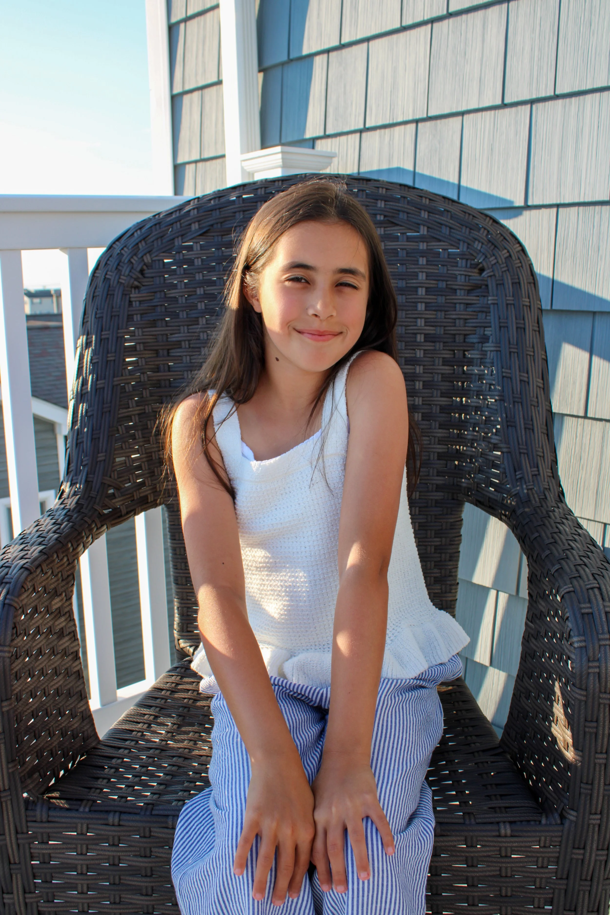 A young girl sitting in a wicker chair on a porch, smiling with squinting eyes, wearing a sleeveless white top and striped blue and white pants, with sunlight casting shadows on her face.