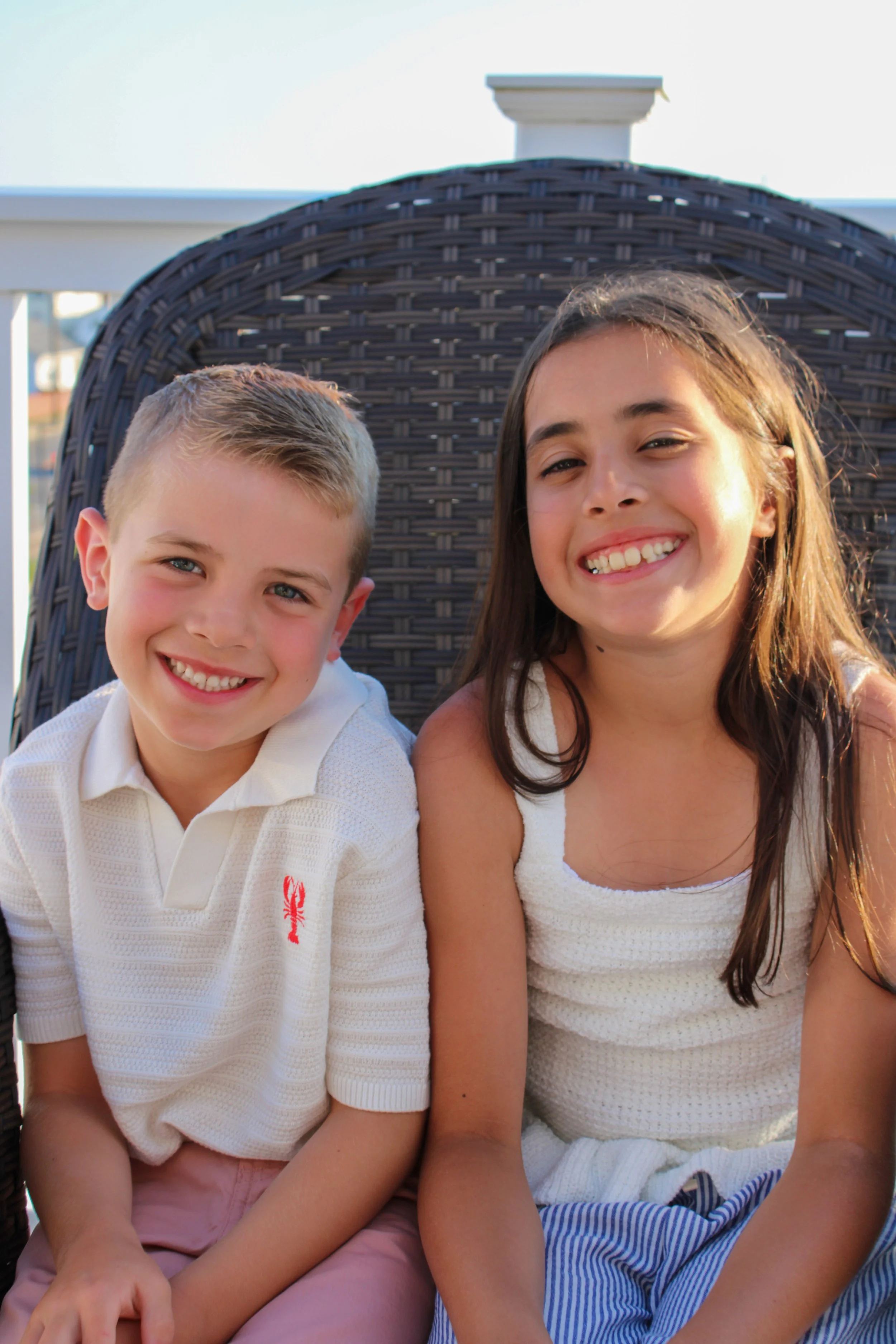 A happy young boy and girl sitting together outdoors, smiling at the camera, with a large wicker chair and part of a white railing in the background.