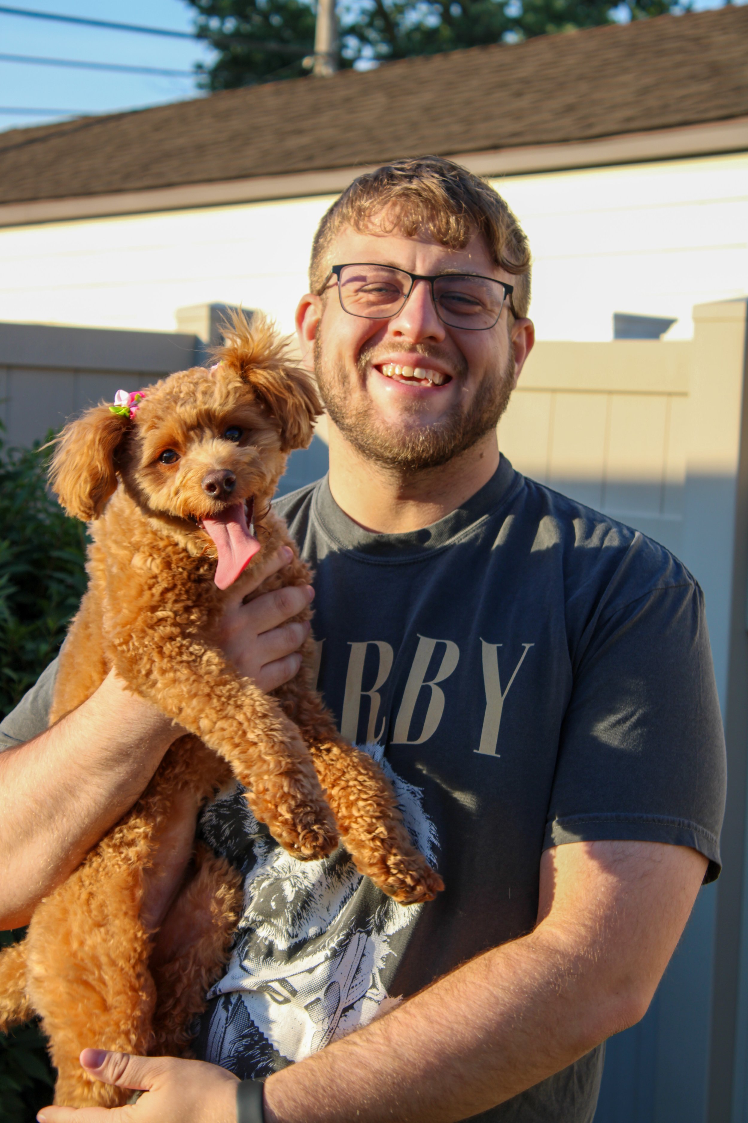 A man wearing glasses and a dark t-shirt holding a small, fluffy brown dog with pink tongue out outside in front of a house with a brown roof and white fence.