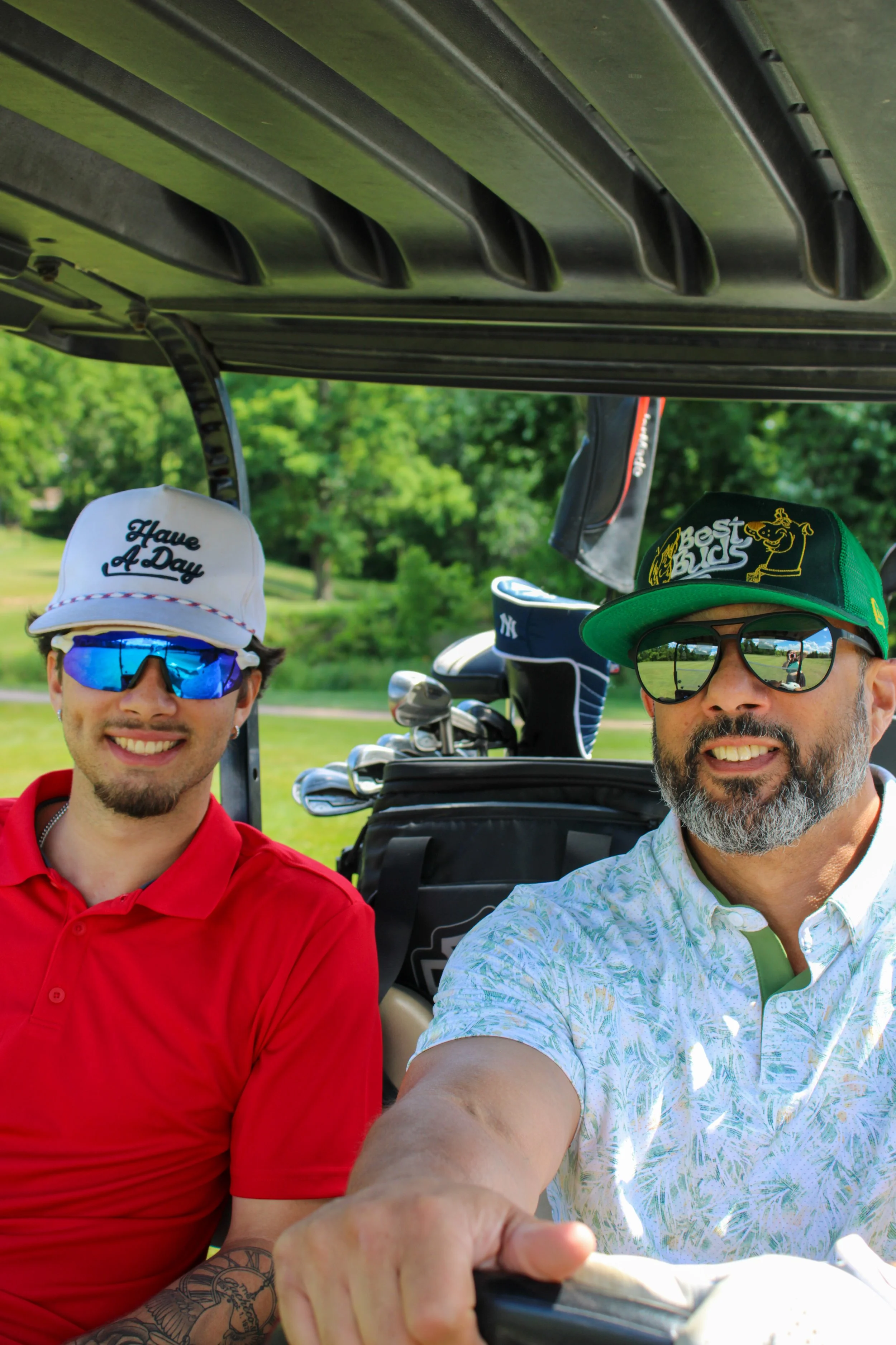Two men in golf carts, both wearing sunglasses and hats, smiling for a selfie on a sunny day with green trees in the background.