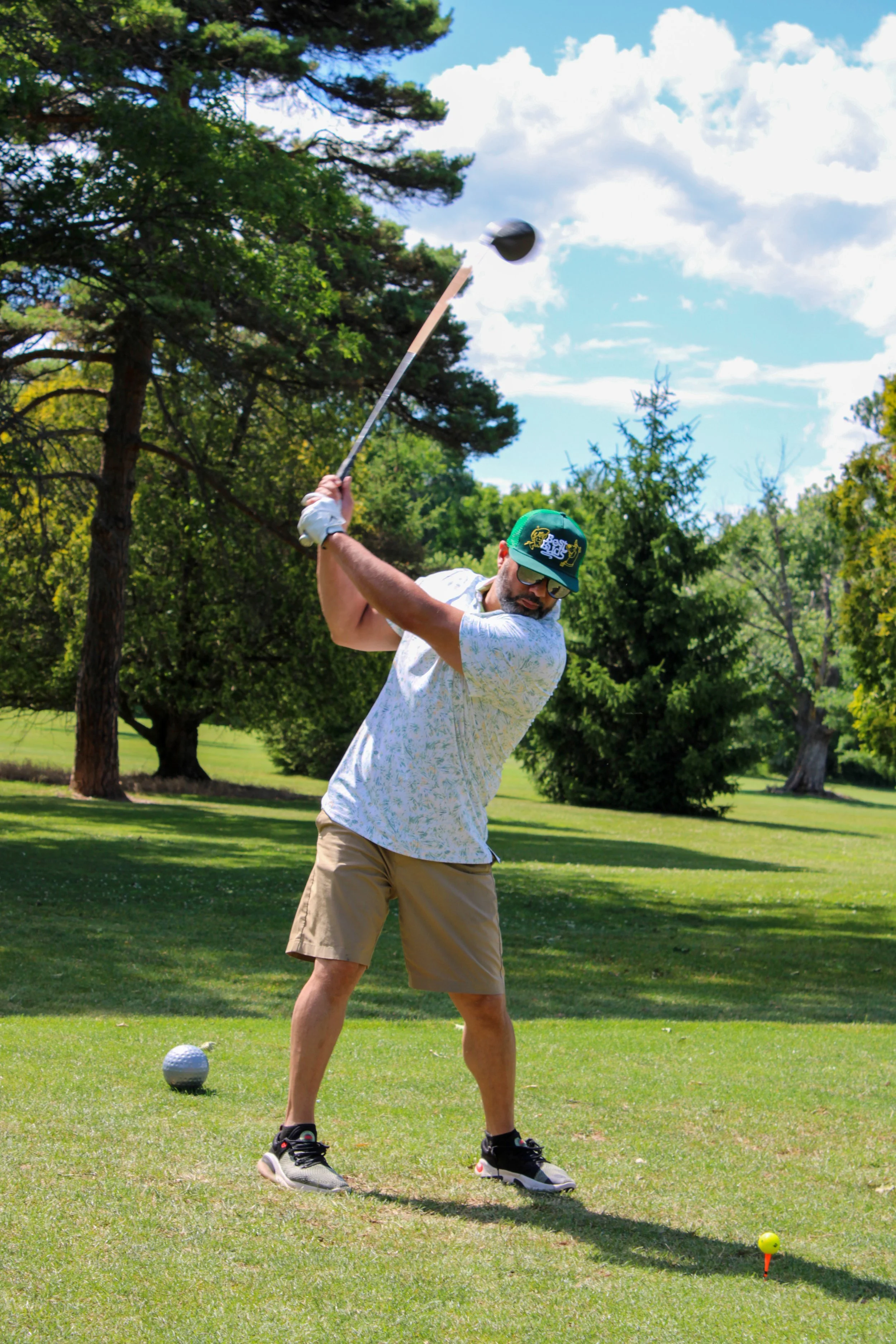 A man playing golf on a lush green golf course, swinging a golf club with a tee and yellow golf ball positioned in front of him, surrounded by trees and a partly cloudy sky.