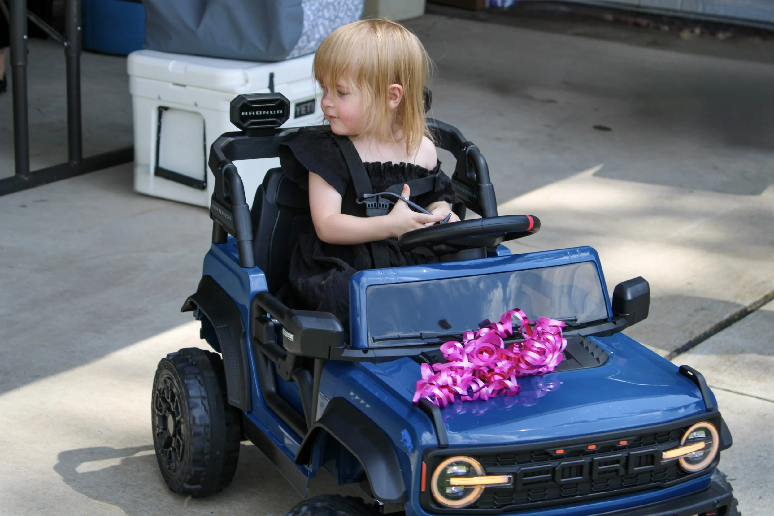 A young girl with blonde hair in a black dress sitting in a blue toy car decorated with pink ribbons, parked outdoors near a garage.