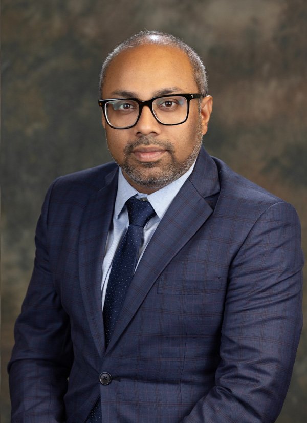 Professional man wearing a navy blue checkered suit, light blue dress shirt, and a dark tie with small white dots, posing against a dark, textured background.