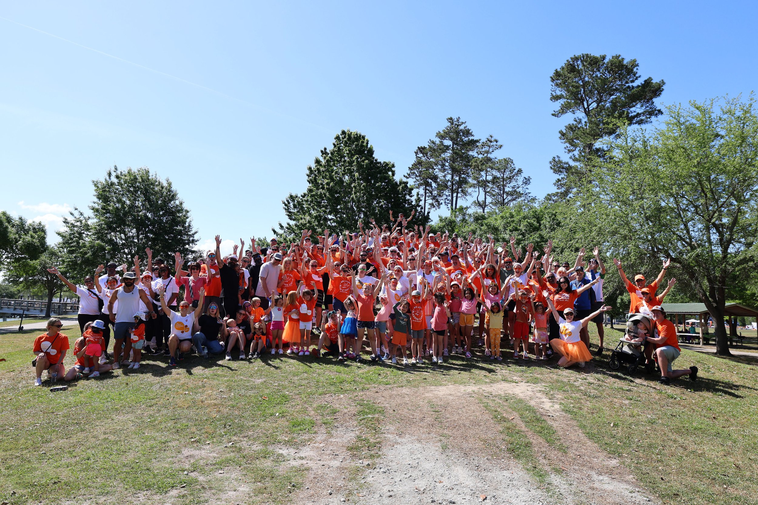A large group of people, including adults and children, gathered outdoors on a sunny day with trees in the background, many wearing orange, pink, and white clothing, some waving. Some individuals are standing, others sitting or crouching, and there is a stroller on the right side.