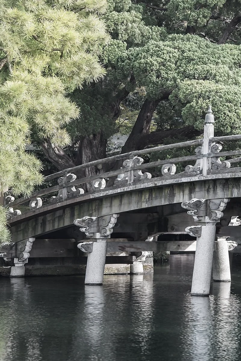 A traditional Japanese stone bridge with decorative railings and support columns, situated over a body of water, surrounded by lush green trees.