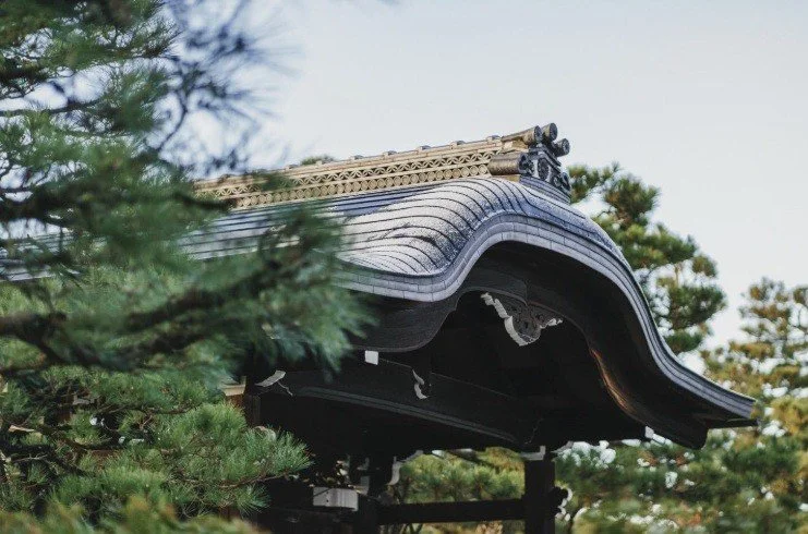 Close-up of a traditional Japanese-style roof with ornate wooden details, surrounded by green trees.