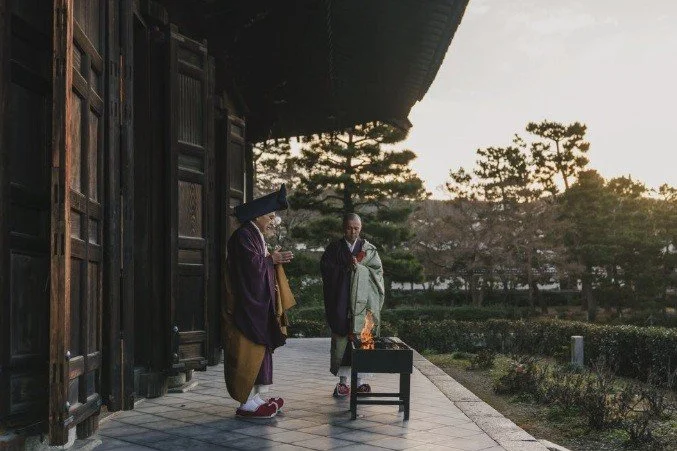 Two monks in traditional robes stand near a small fire pit outside a temple, with trees and a pathway in the background at sunset.