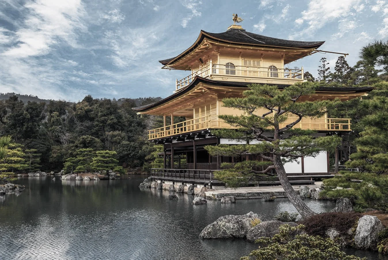 Traditional Japanese building with multiple levels beside a pond with rocks and greenery, under a partly cloudy sky.