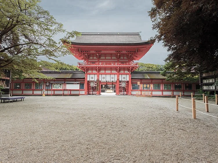 Traditional Japanese temple gate with red and white wood structure, surrounded by trees and a gravel courtyard.
