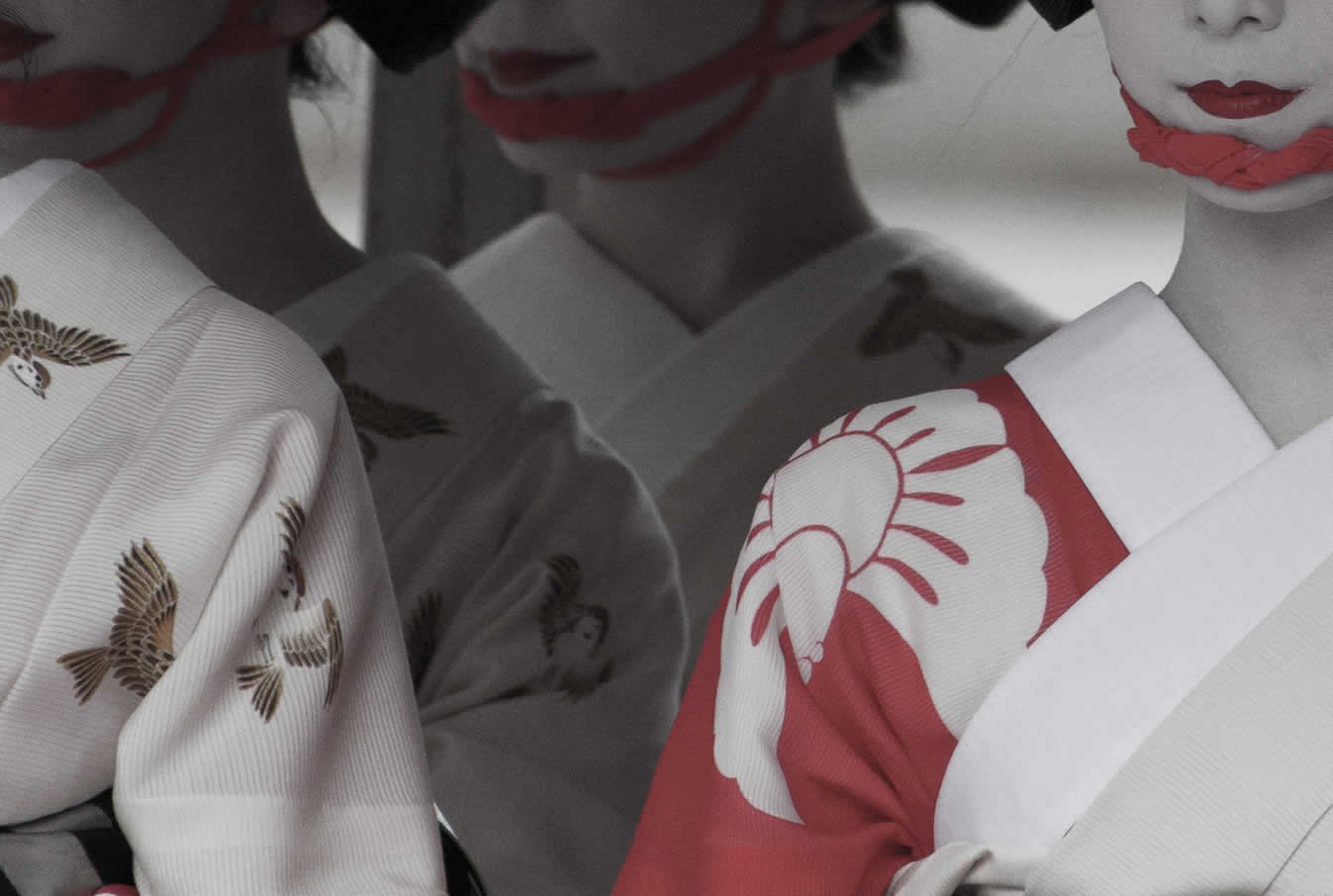 Close-up of three Japanese women wearing traditional kimonos and white makeup with red lips, with the focus on one woman’s shoulder and face, while the others are in the background.