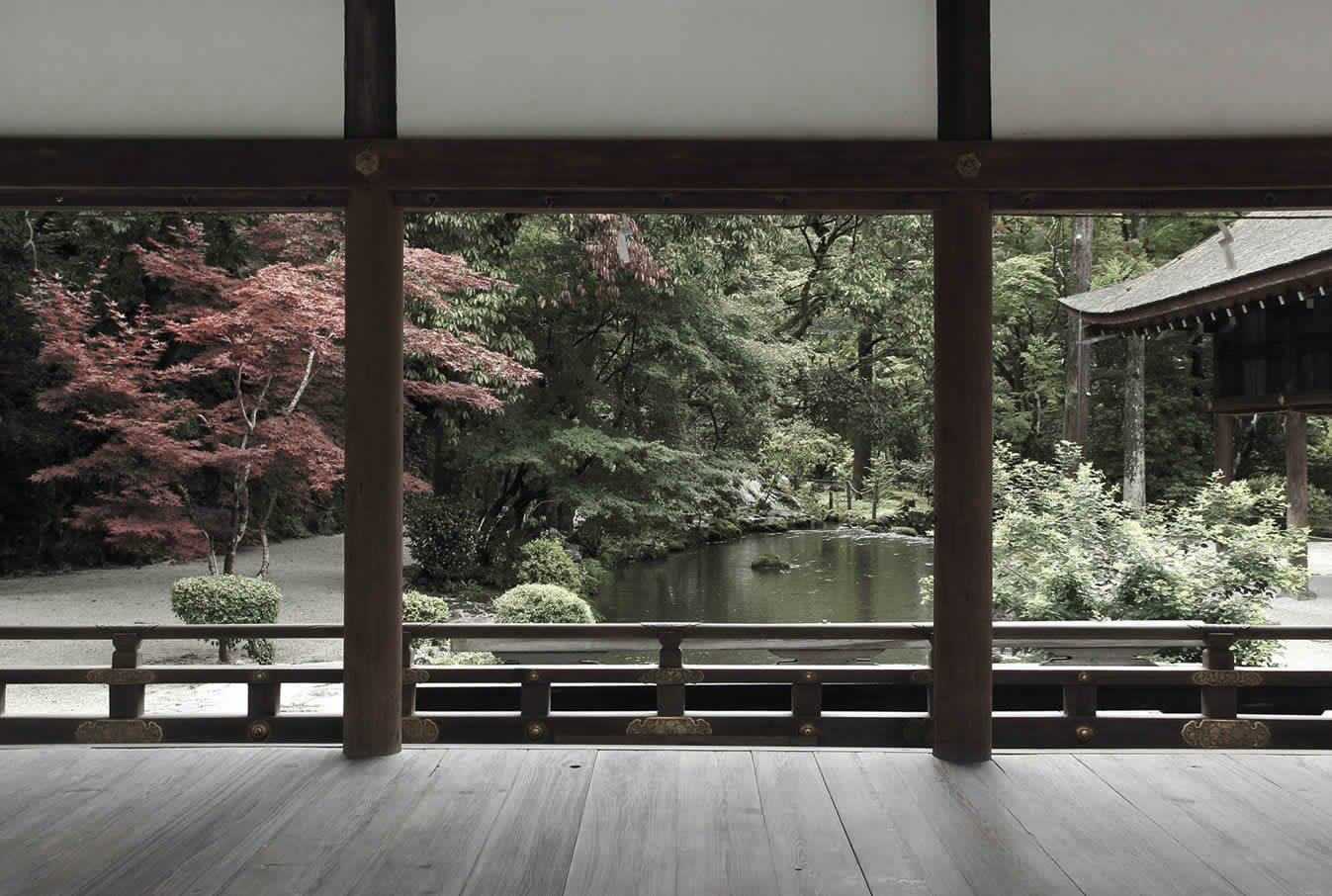 View from a traditional Japanese wooden temple or pavilion showing a serene garden with a pond, trees, bushes, and a small building in the background.