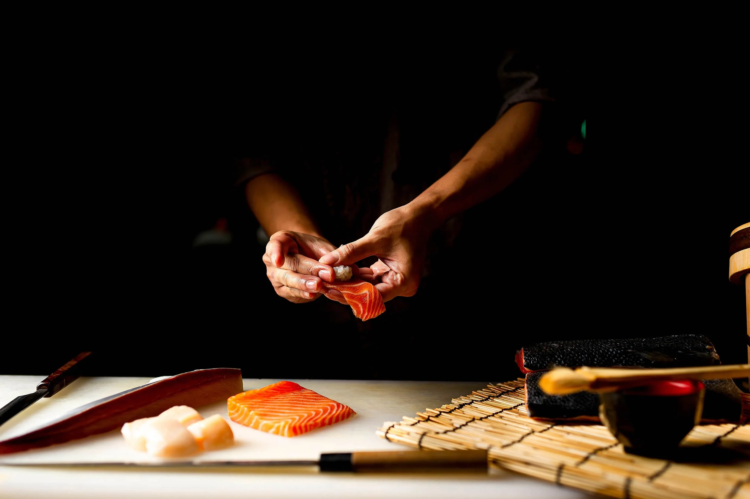 A person preparing sushi with various fish slices on a cutting board, using a bamboo sushi rolling mat, with a dark background.