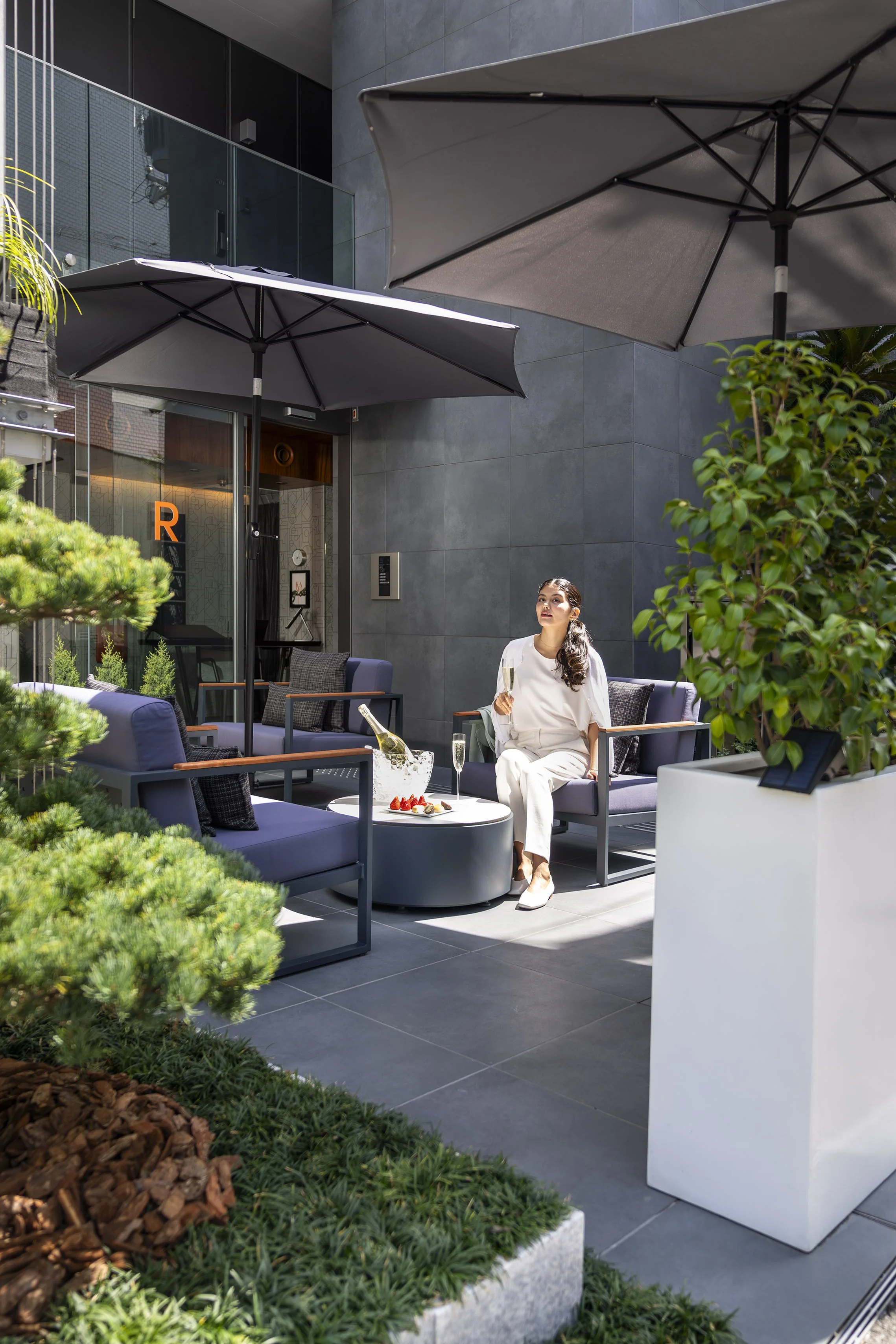 A woman sitting outdoors on a patio, holding a glass of champagne, with a table of snacks in front of her. The patio has large umbrellas, modern furniture, and is surrounded by greenery, with a modern building in the background.