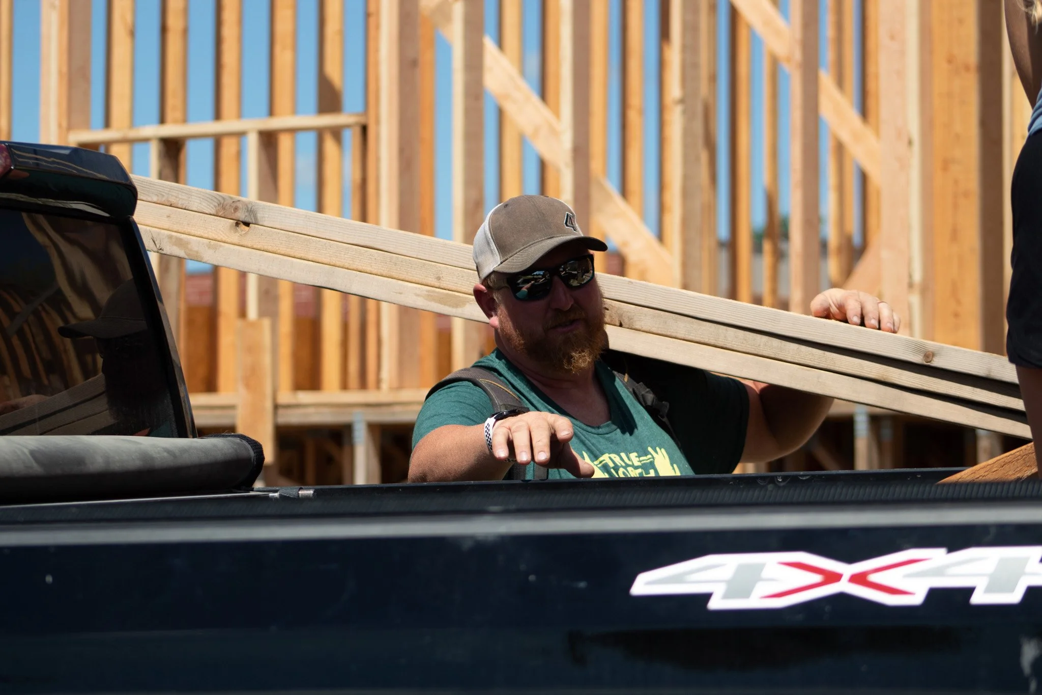 Man wearing sunglasses, baseball cap, and a green shirt with yellow text, carrying wooden planks at a construction site with a background of unfinished wooden frame and blue sky.
