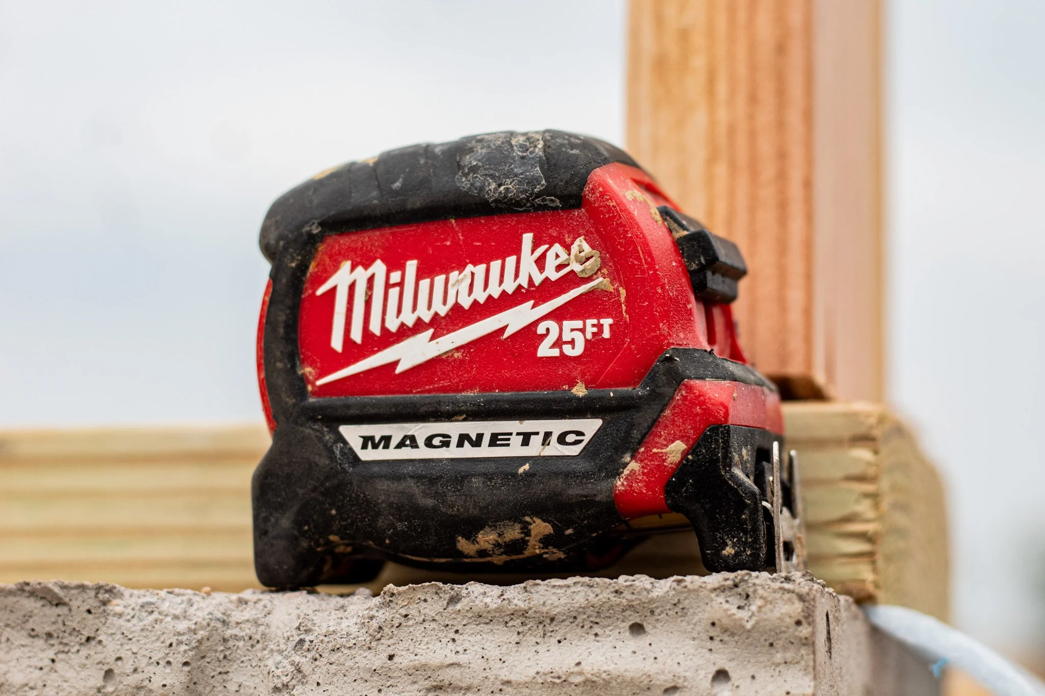 Red Milwaukee cordless drill with black and red grip resting on a concrete block. The drill shows signs of use, with visible dirt and wear, and has a white sticker that says 'MAGNETIC'. The background is blurred but includes a wooden piece of framing and a white wall.