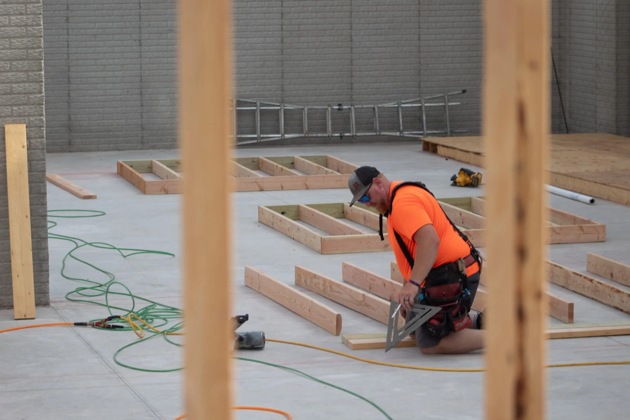 A construction worker wearing an orange shirt, cap, and sunglasses working on framing a building interior. Wooden frameworks are on the concrete floor, with tools and green extension cords scattered around.