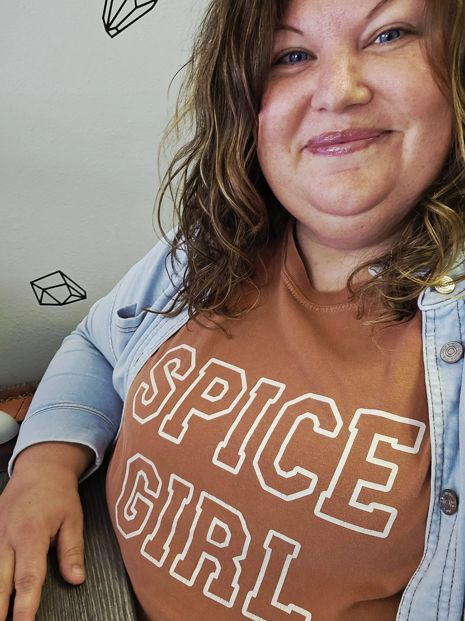 A woman with shoulder-length wavy hair, smiling slightly, wearing a orange t-shirt with large white outline text reading 'SPICE' and a light denim jacket, seated with her arm resting on a wooden table.
