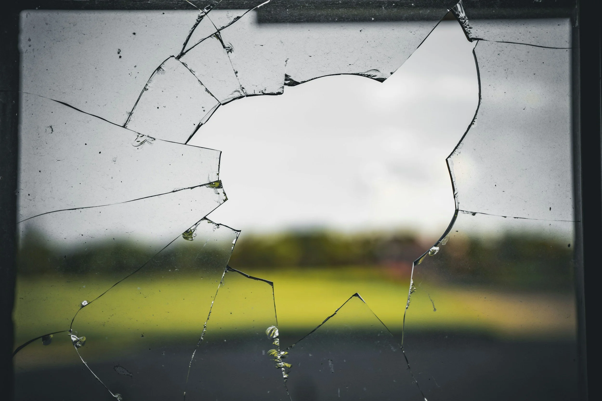 Broken glass window with multiple cracks and missing pieces, showing a blurred outdoor background with green grass and trees.