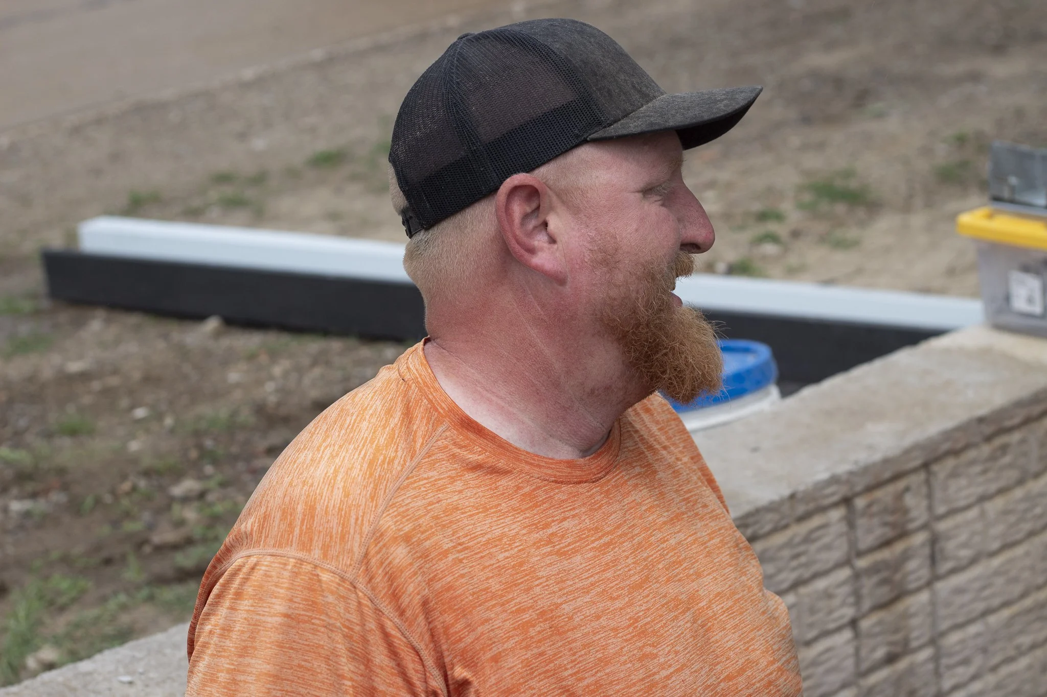 A man with a red beard and a beige skin tone, wearing an orange shirt and a black snapback cap, is in profile view outdoors.
