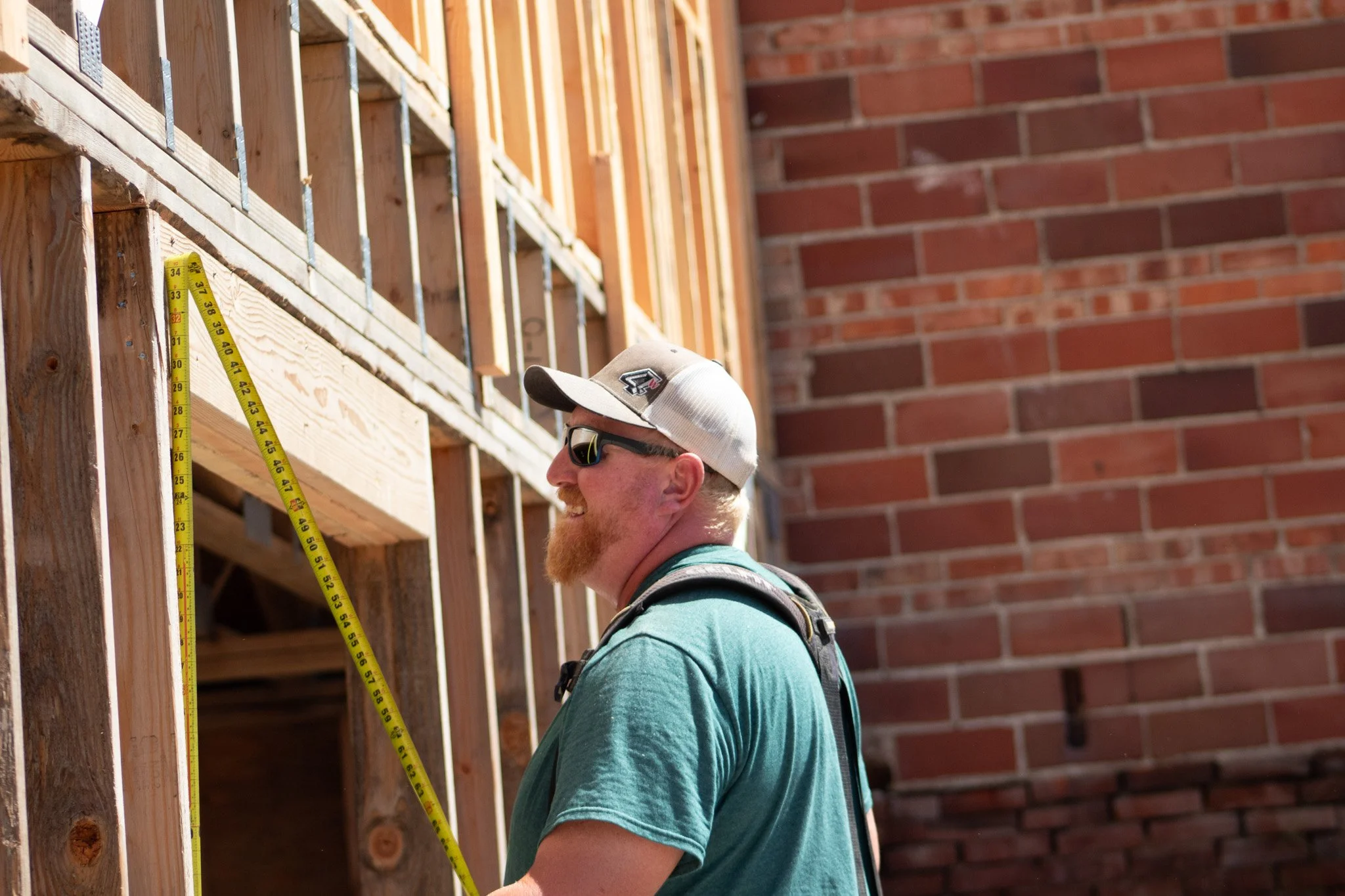 A construction worker wearing sunglasses, a white cap, and a teal shirt is looking at a wooden wall with measuring tapes attached to the wall, standing in front of a brick wall.